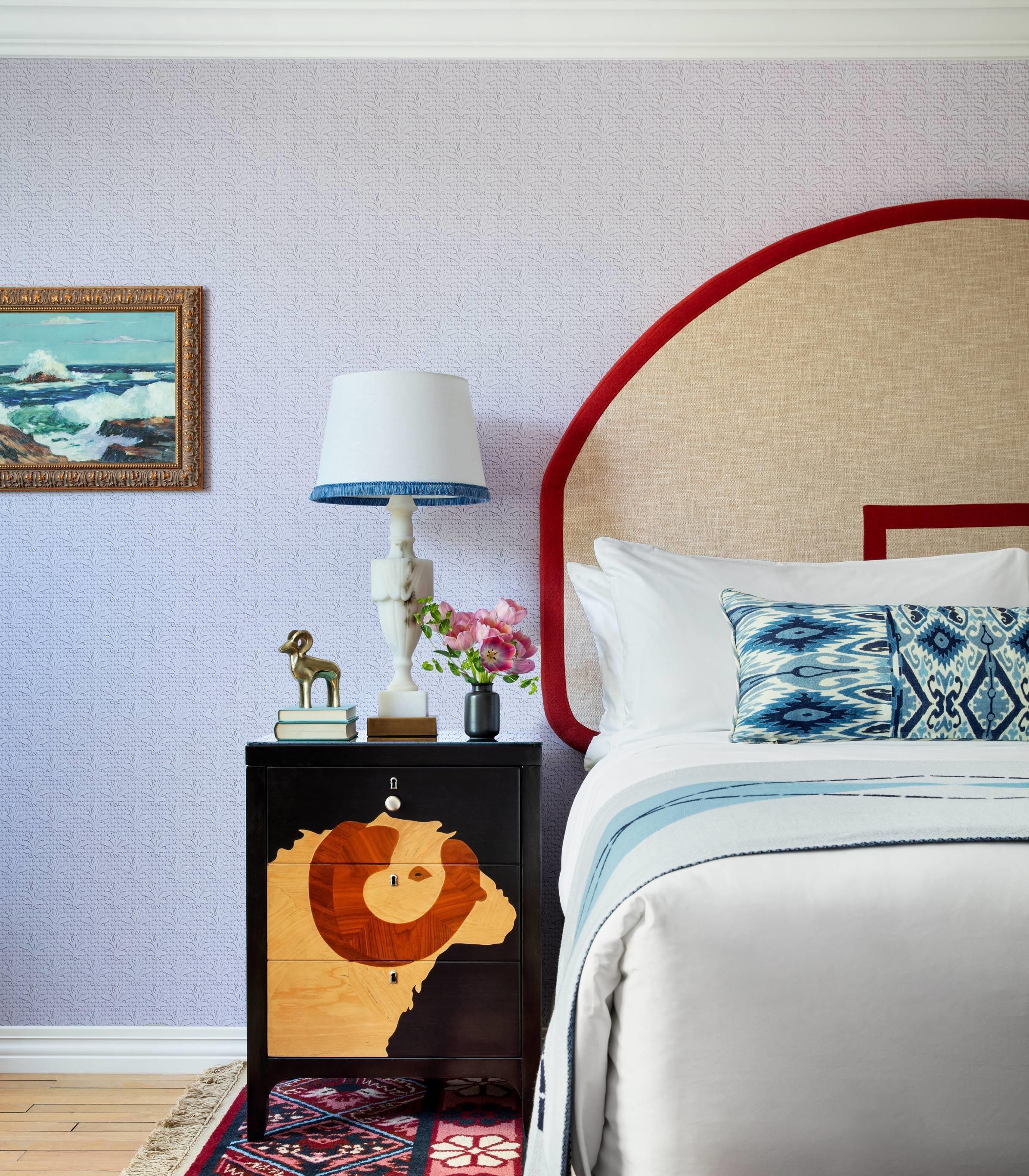 A bedroom with a bed, a nightstand with a lamp, a framed painting, and a decorative wooden piece.