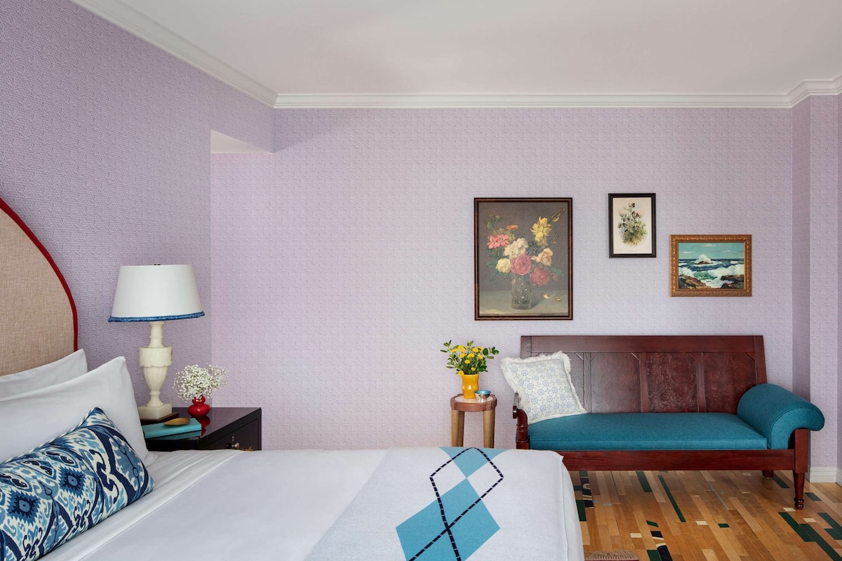 A hotel room with a bed, a sofa, a nightstand with a lamp, and two framed pictures on the wall.