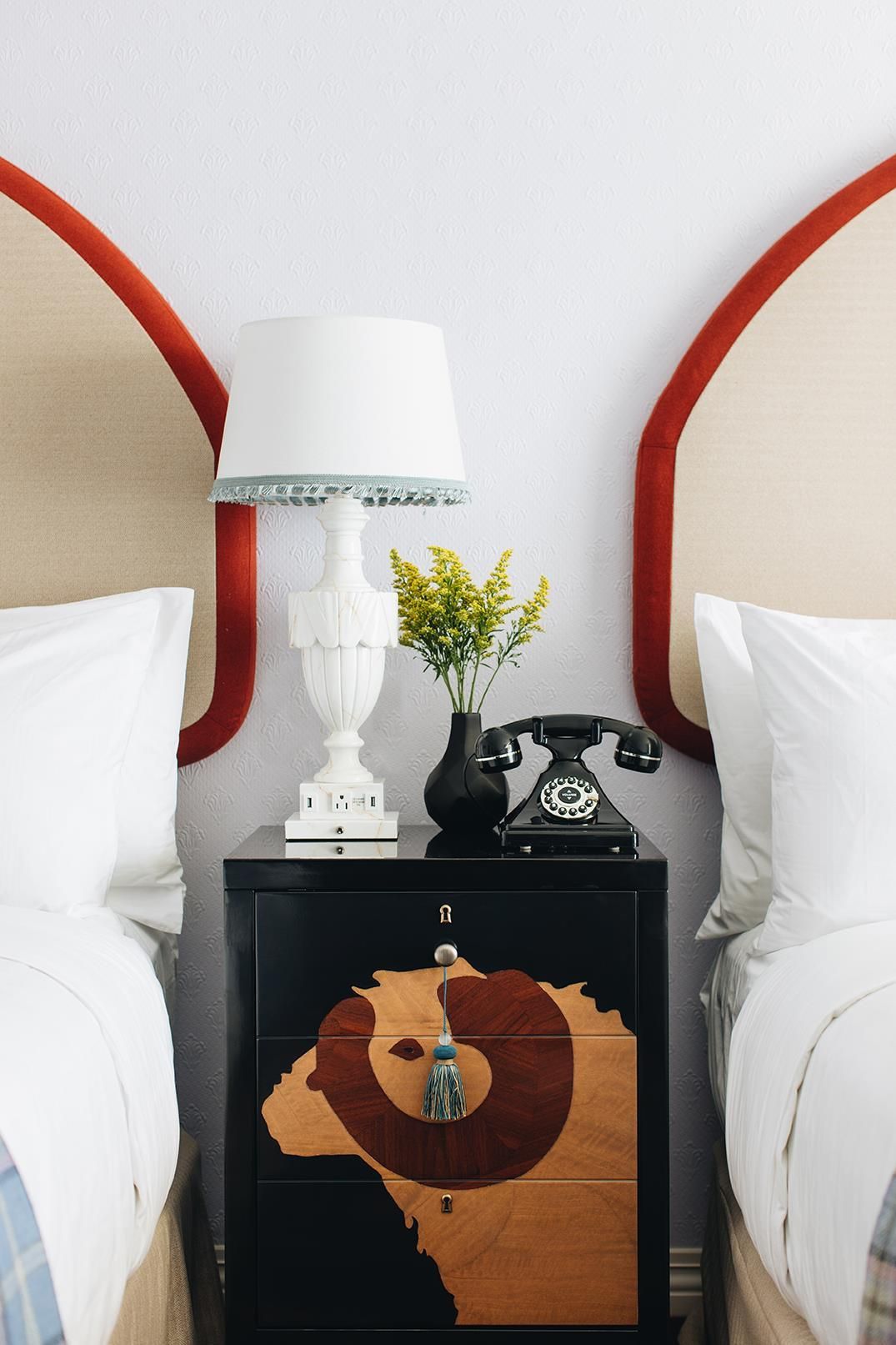 Un lit bien fait, avec du linge de lit blanc, une table de chevet avec une lampe, un vase rempli de fleurs et un vieux téléphone à cadran.