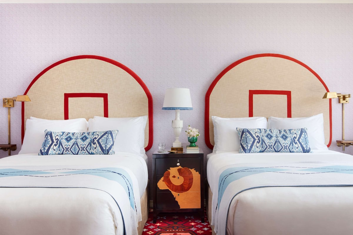 A hotel room with two beds, each with a red headboard, white bedding, and patterned pillows. There is a nightstand between the beds with a lamp and a vase of flowers.