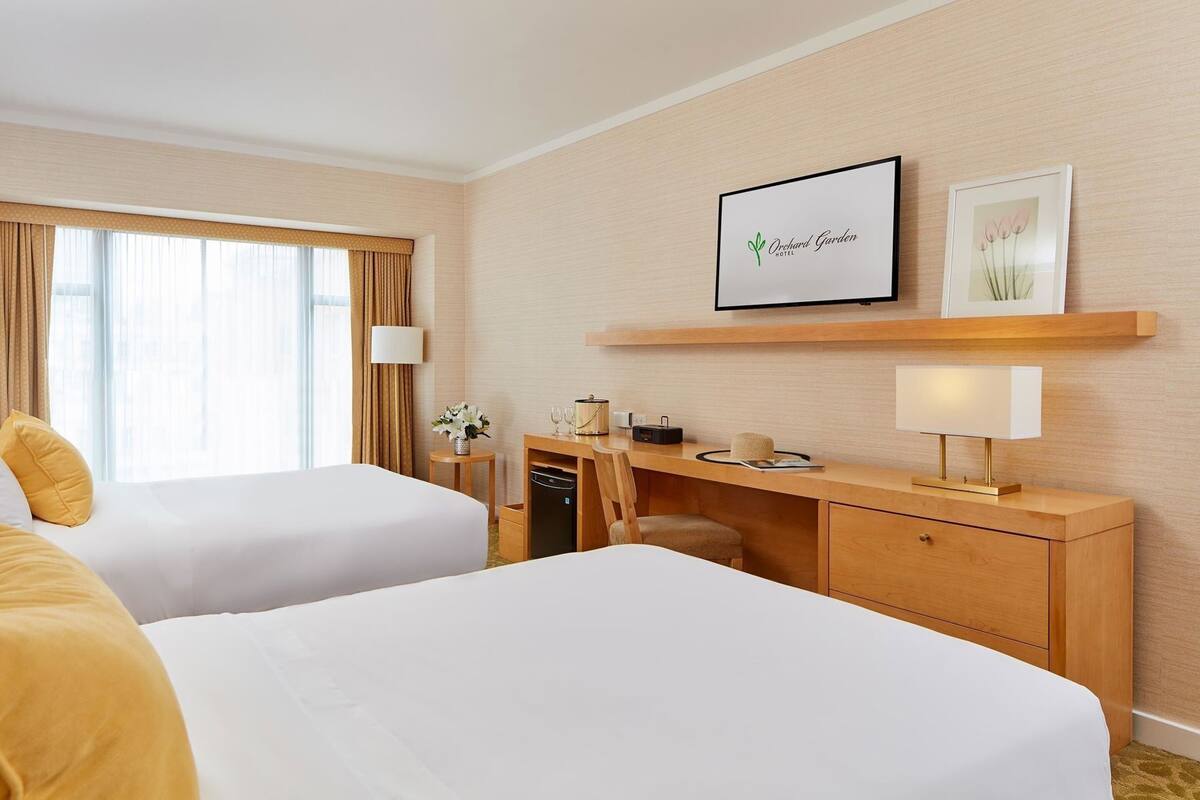 A hotel room with two beds, a desk, a lamp, a framed picture, and a window with curtains.