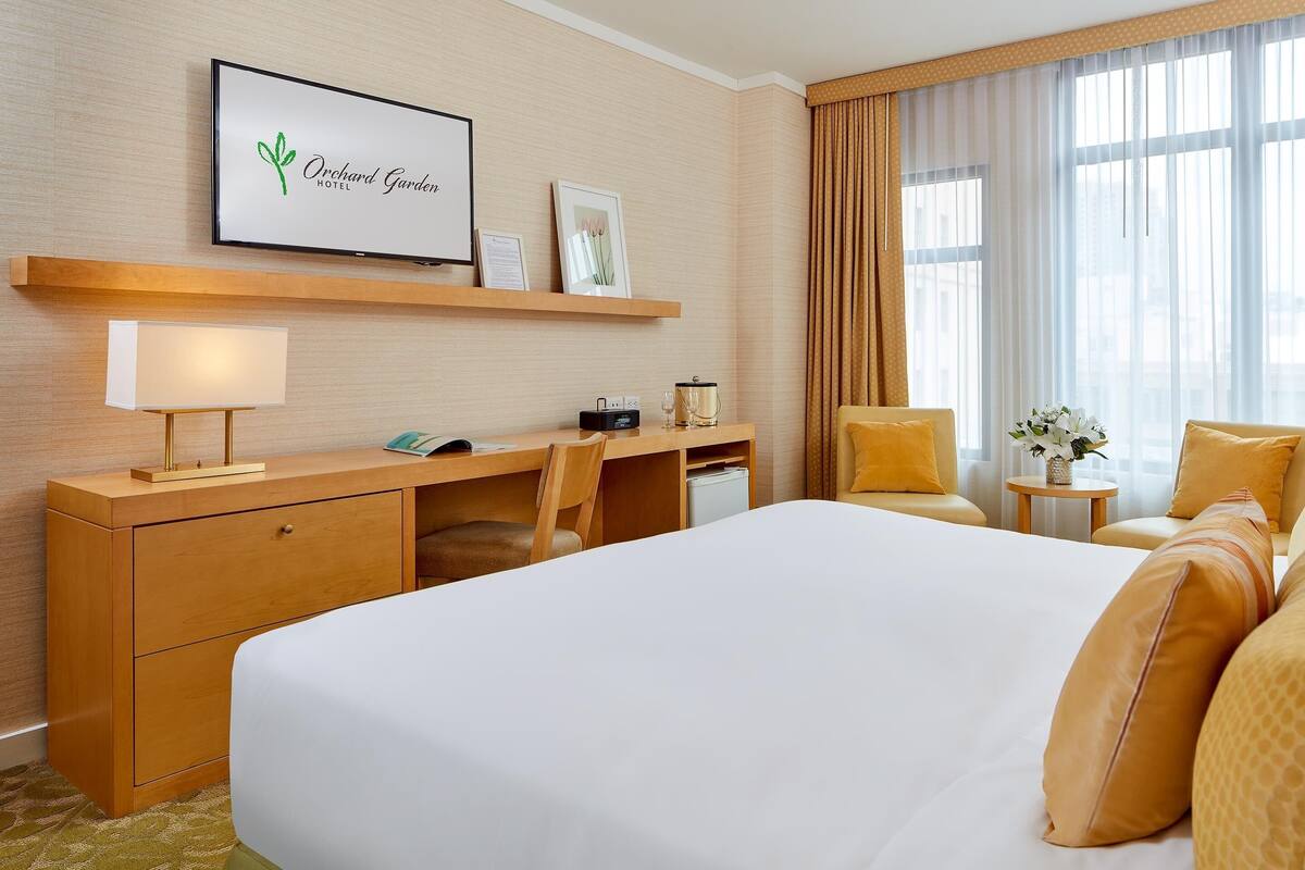 A hotel room with a large bed, a desk with a lamp, a chair, a small table with a vase, and a large window with curtains.