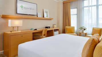 A hotel room with a large bed, a desk with a lamp, a chair, a small table with a vase, and a large window with curtains.