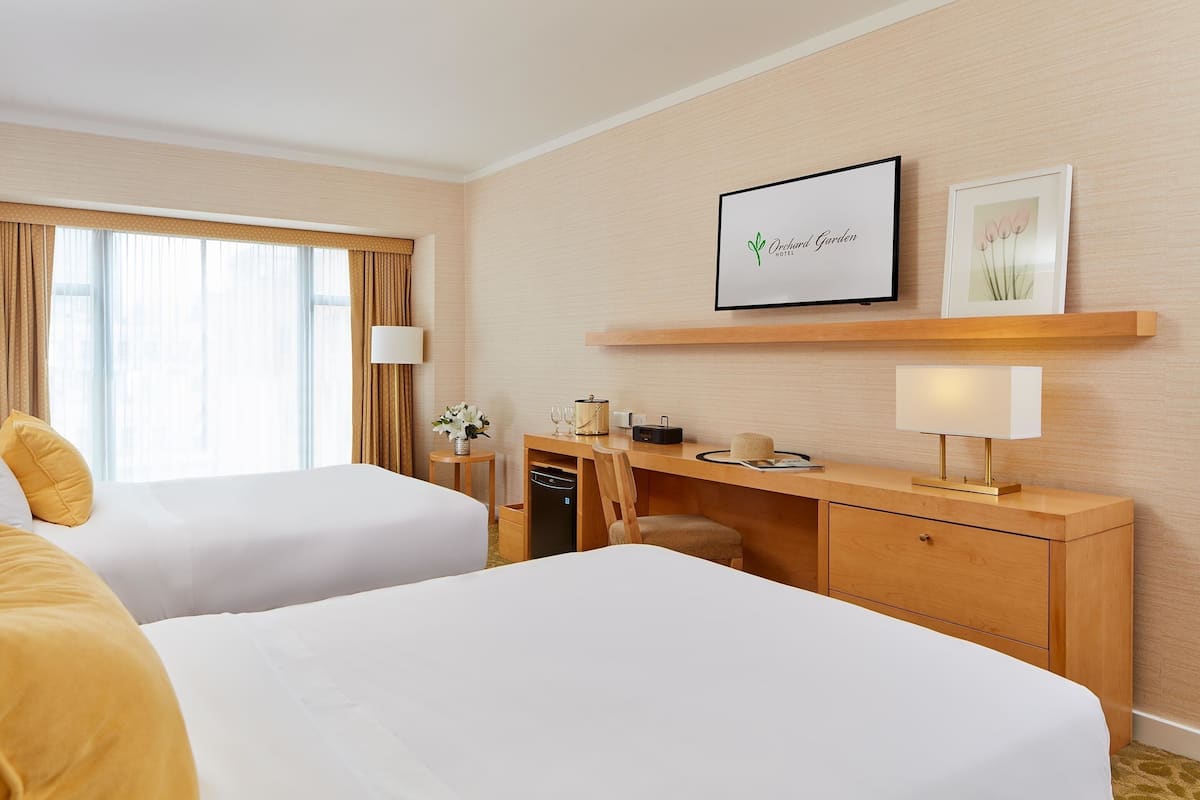 A hotel room with two beds, a desk with a lamp, a small table with a vase, and a framed picture on the wall.