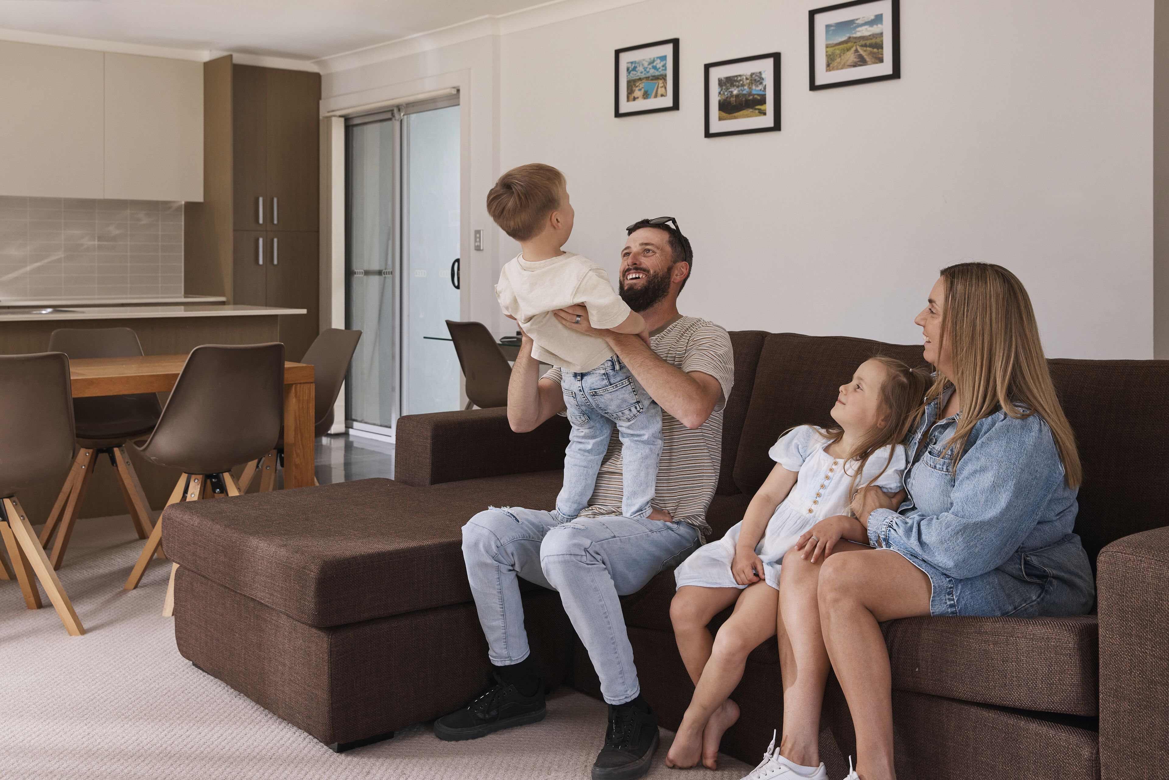 A family sitting on a brown sectional sofa in a modern living room with a dining area and kitchen in the background.