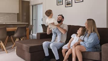 Une famille est assise sur un canapé d’angle marron dans un salon moderne, avec un coin repas et une cuisine en arrière-plan.