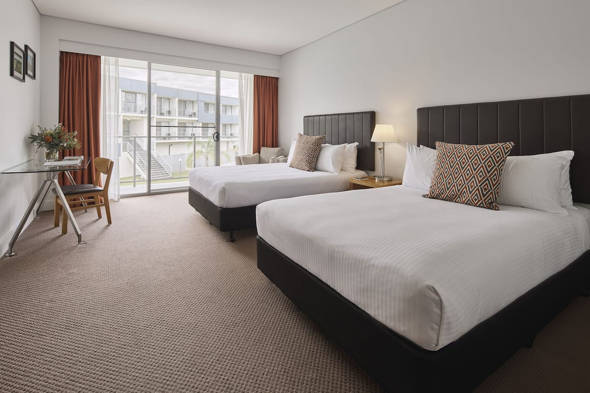 A hotel room with two beds, a desk with a chair, a window with curtains, and a view of the building outside.