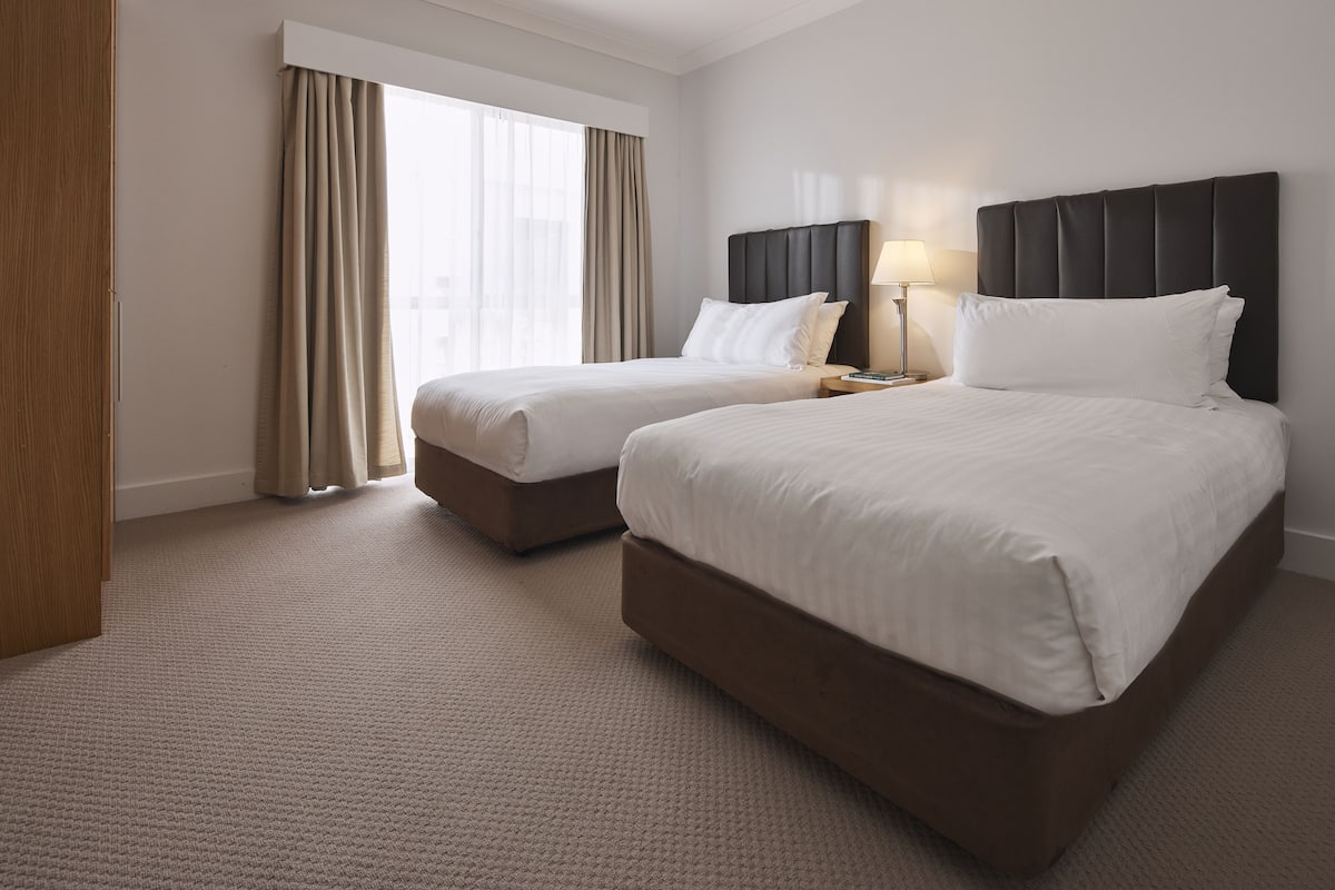 A hotel room with two beds, a large window with curtains, a bedside table with a lamp, and a carpeted floor.