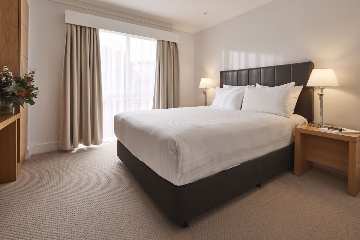 A hotel room with a large bed, two bedside tables with lamps, a wooden nightstand, and a large window with curtains.