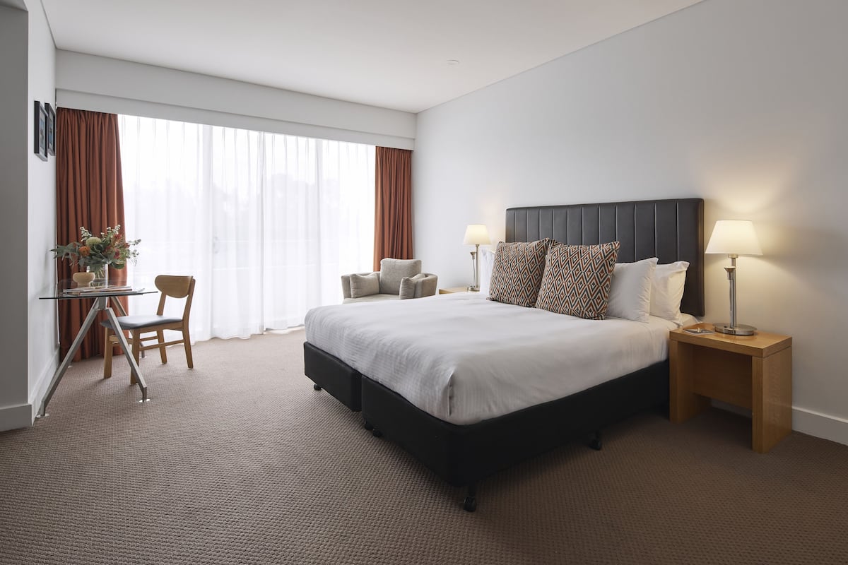 A hotel room with a large bed, two bedside tables with lamps, a small desk with a chair, and a large window with curtains.