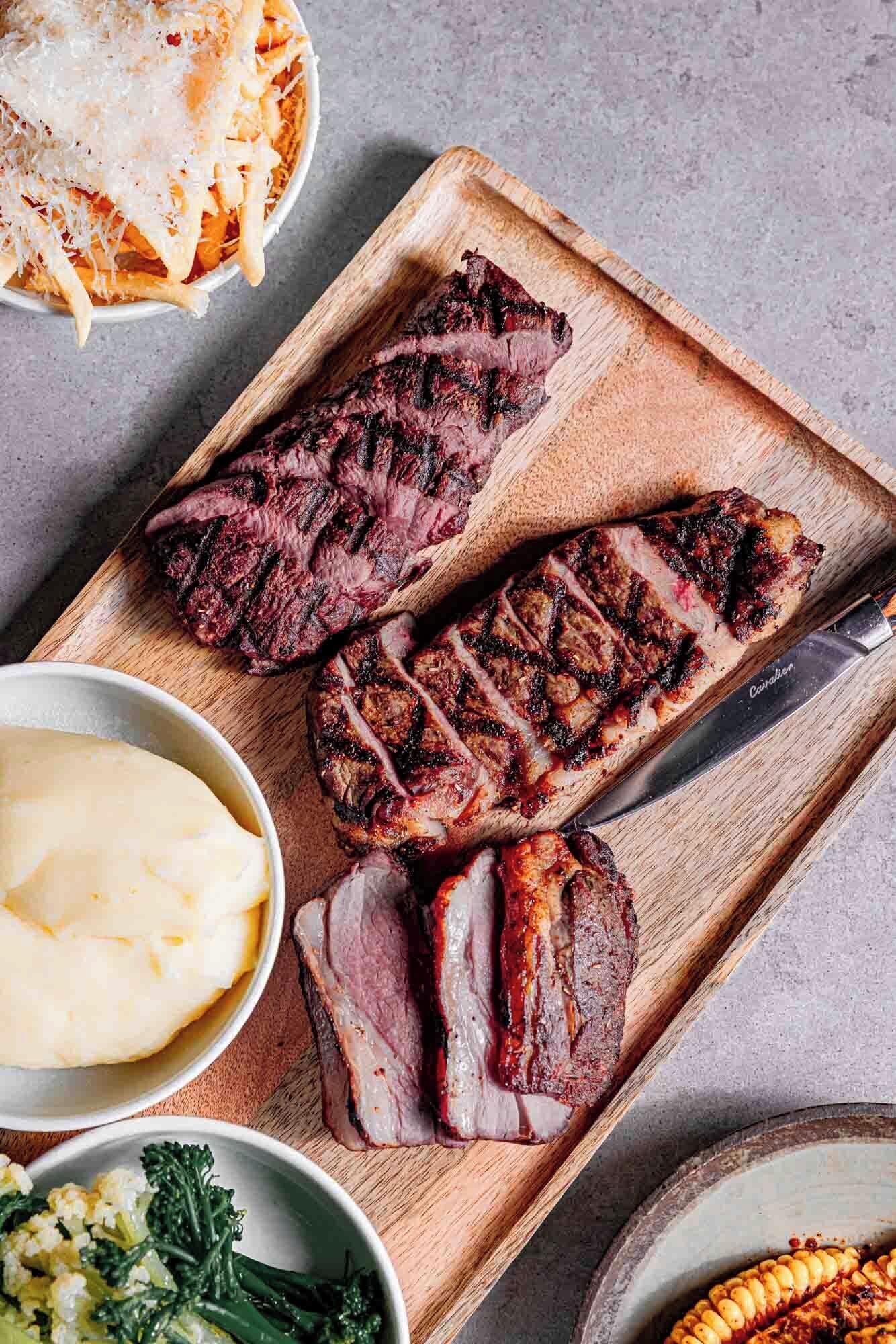 Une planche en bois sur laquelle sont servis un steak grillé, de la purée de pommes de terre et un accompagnement de légumes.