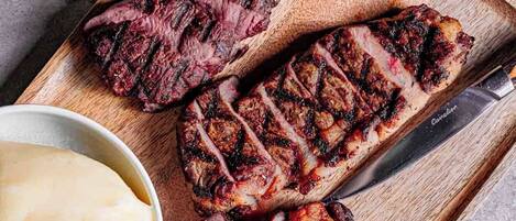 Une planche en bois sur laquelle sont servis un steak grillé, de la purée de pommes de terre et un accompagnement de légumes.