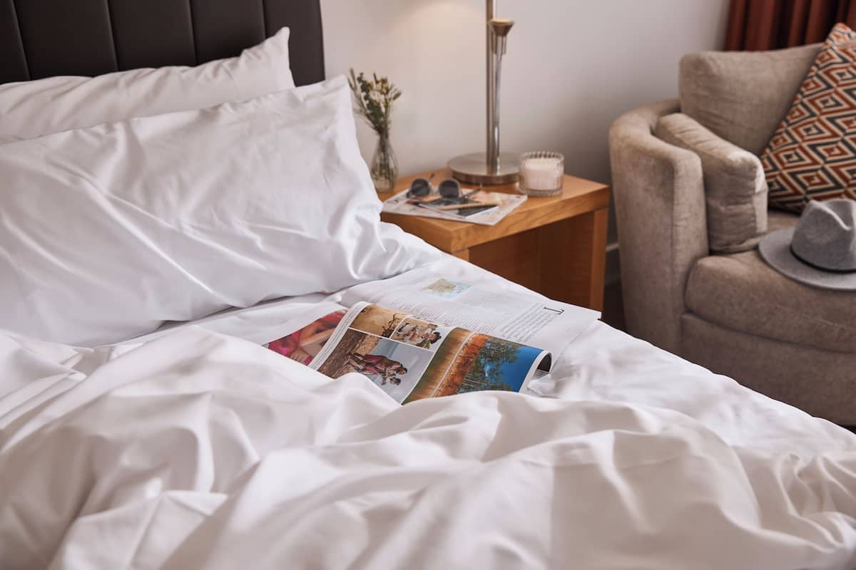 A neatly made bed with white linens, a magazine on the bed, a bedside table with a lamp, and a chair with a patterned cushion.