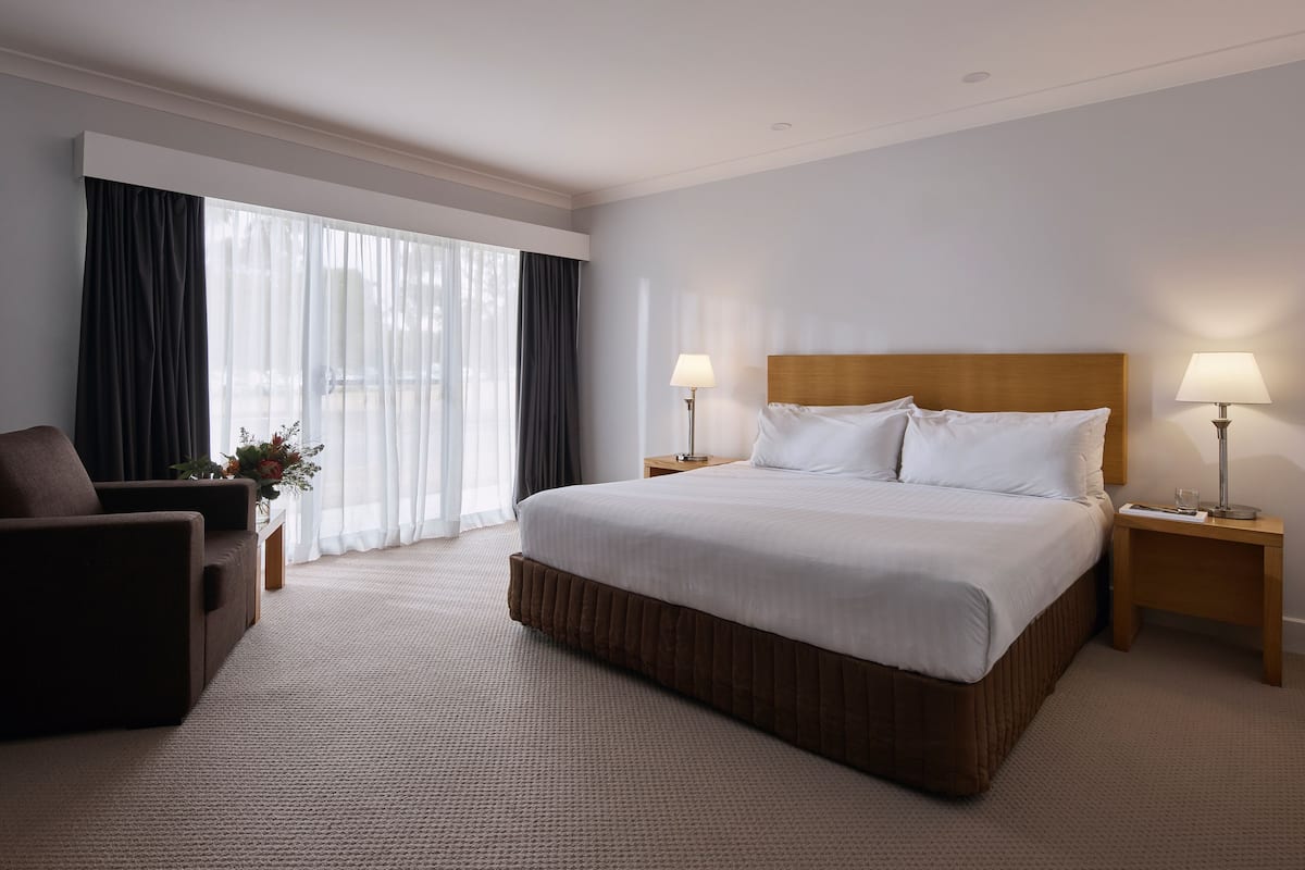 A hotel room with a large bed, two bedside tables with lamps, a dark armchair, and a window with sheer curtains.