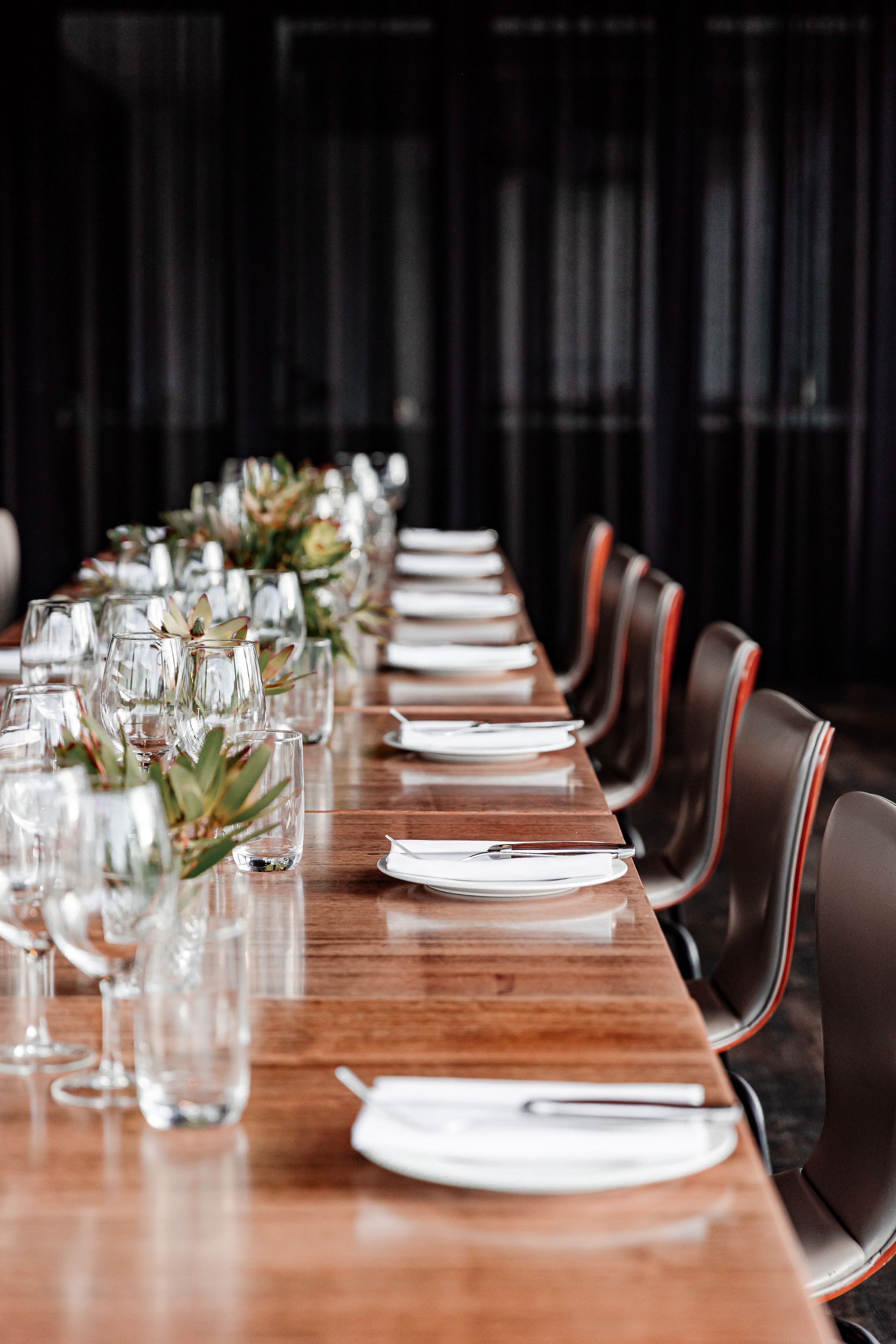 A long wooden table set with clear glasses, white plates, and silverware, flanked by leather chairs with red stripes.
