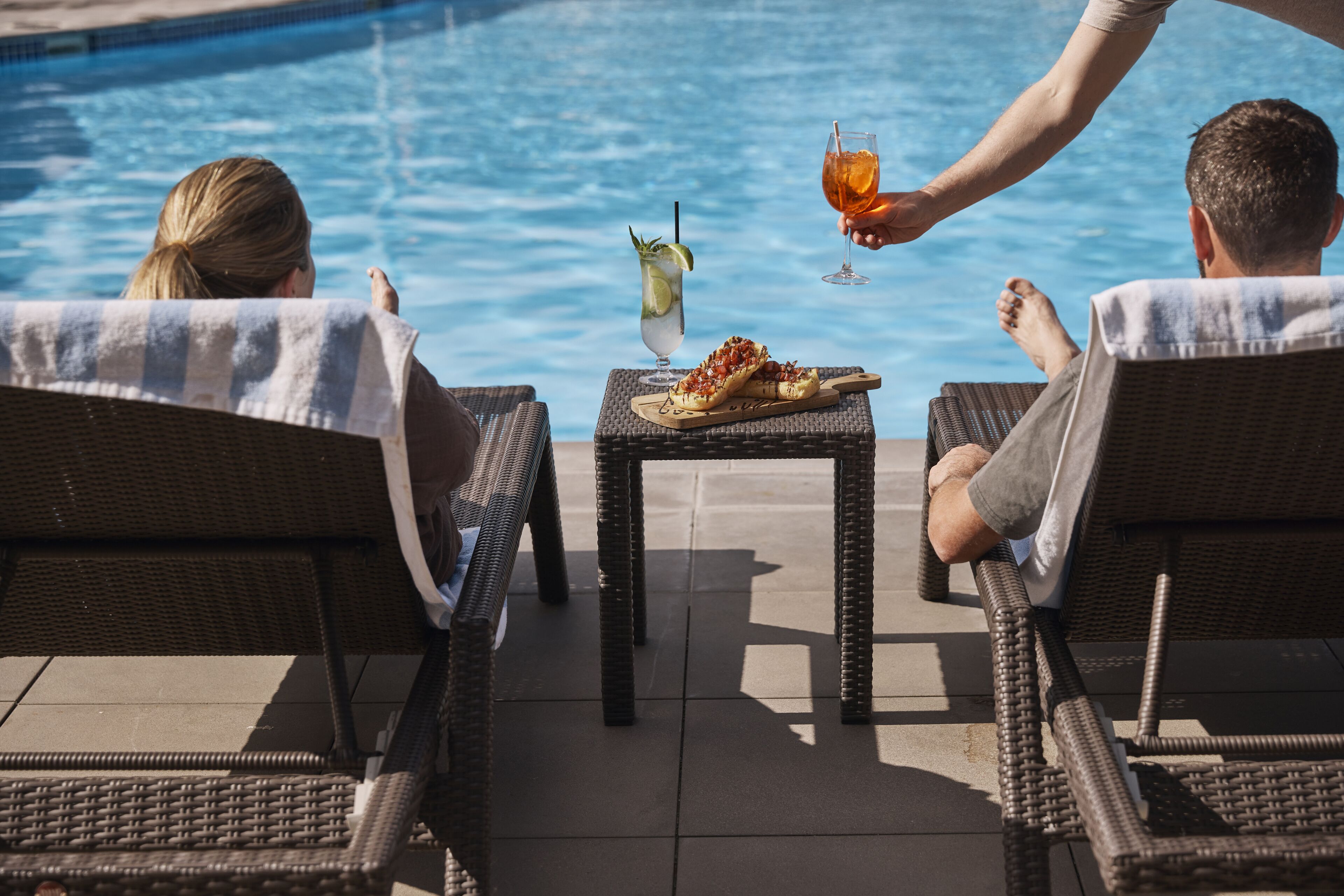 Un área junto a la piscina con sillones de mimbre, una mesita con bebidas y aperitivos, y una vista despejada de la piscina.