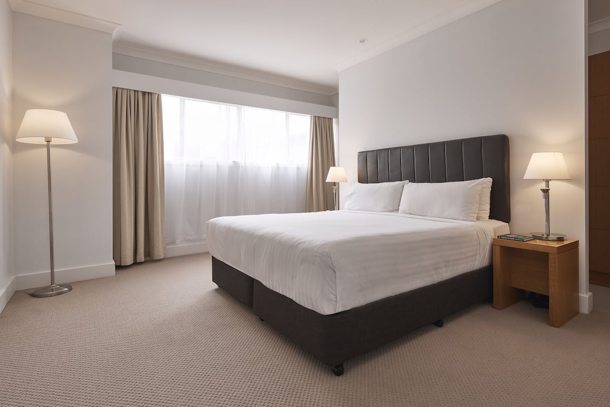 A hotel room with a large bed, two bedside lamps, a window with curtains, and a carpeted floor.