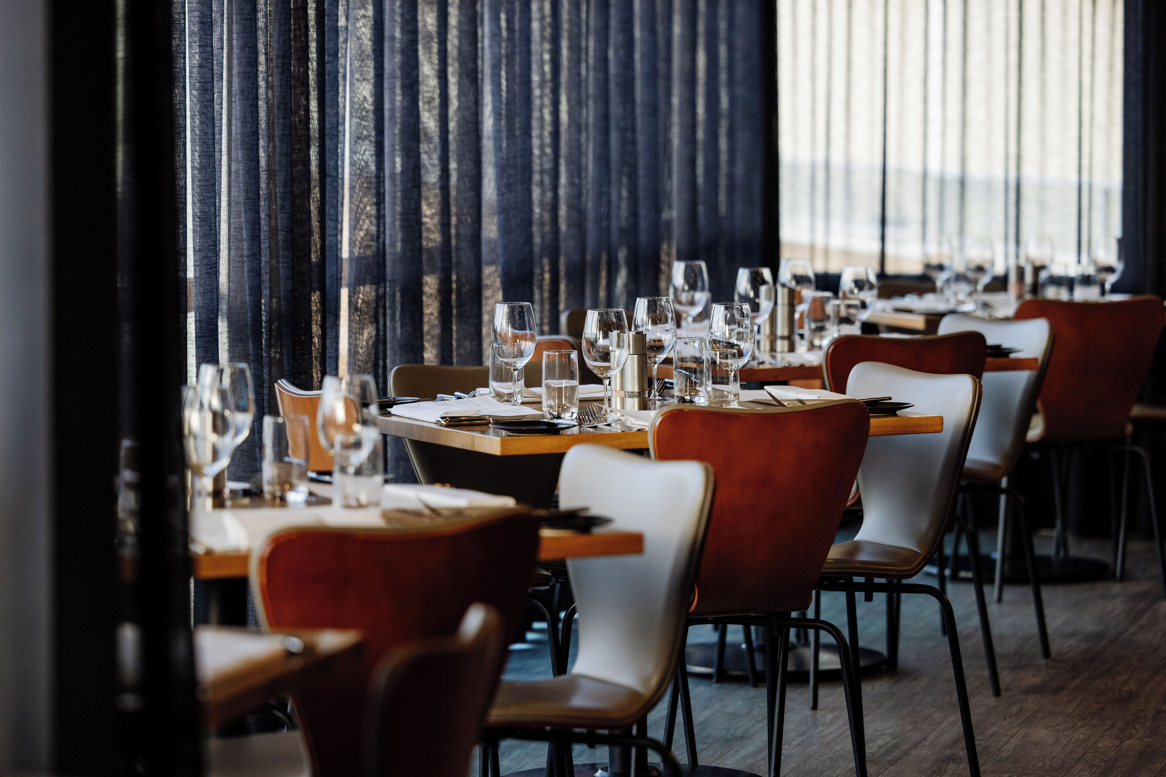 A restaurant with a long table set for dining, chairs, and a view through large windows.