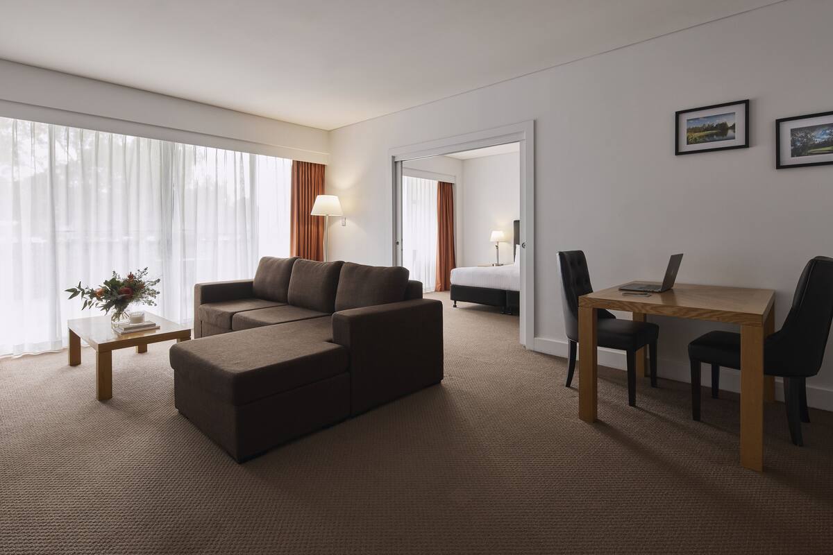 A hotel room with a brown sofa, a small table with a lamp, a wooden desk with a laptop, and a bed in the background.