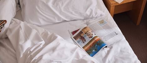 A neatly made bed with white linens, a magazine on the bed, and a wooden nightstand with a lamp and a glass container.