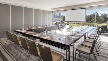 A conference room with a long table, chairs, a projector screen, and a large window overlooking a pool and outdoor area.