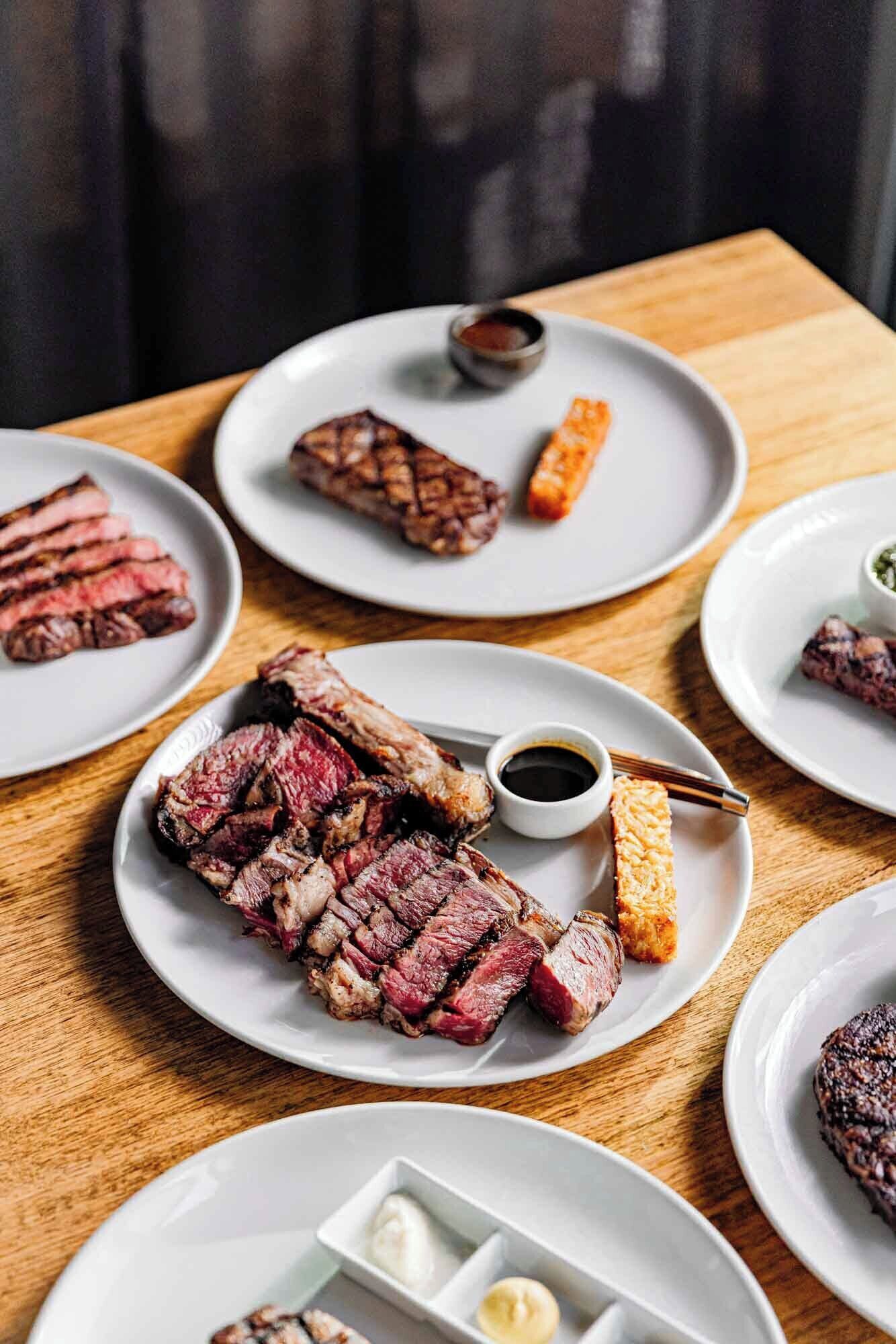 A table with multiple plates of steak, each accompanied by condiments.