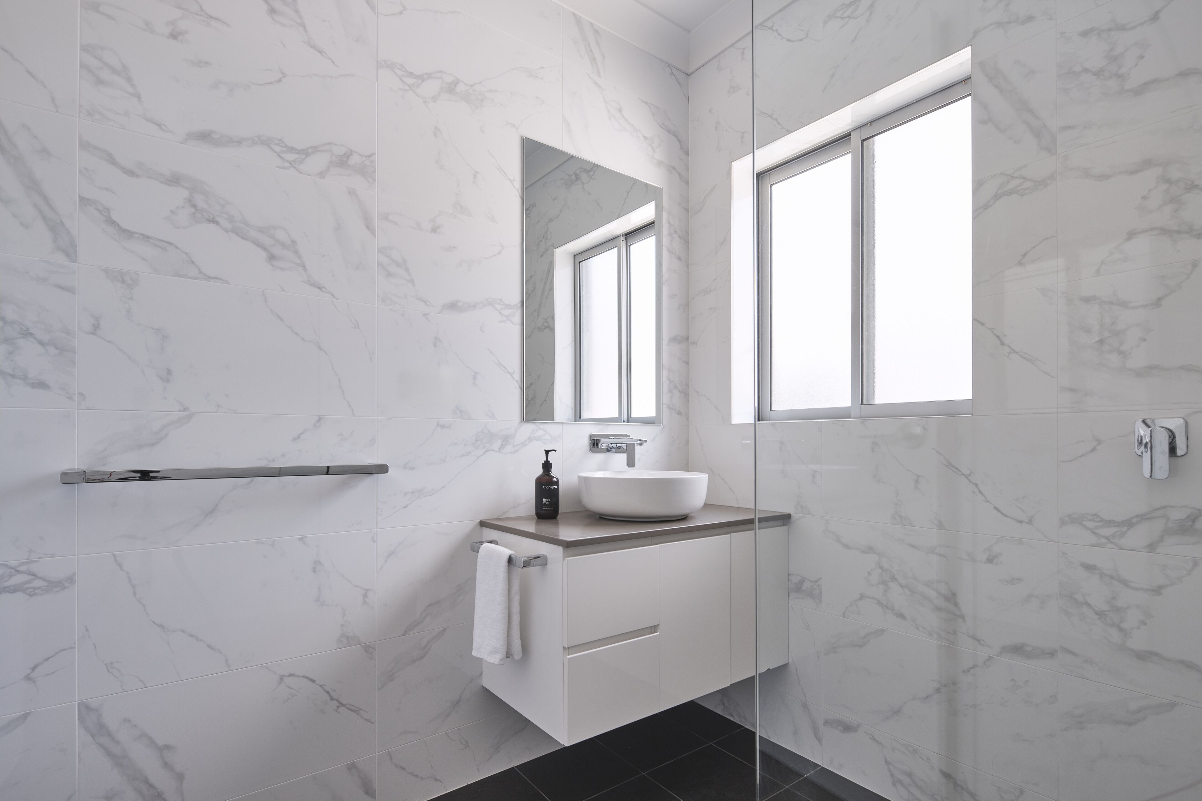 Un baño moderno con pared de mármol, un espejo grande, un lavamanos blanco y una ventana.