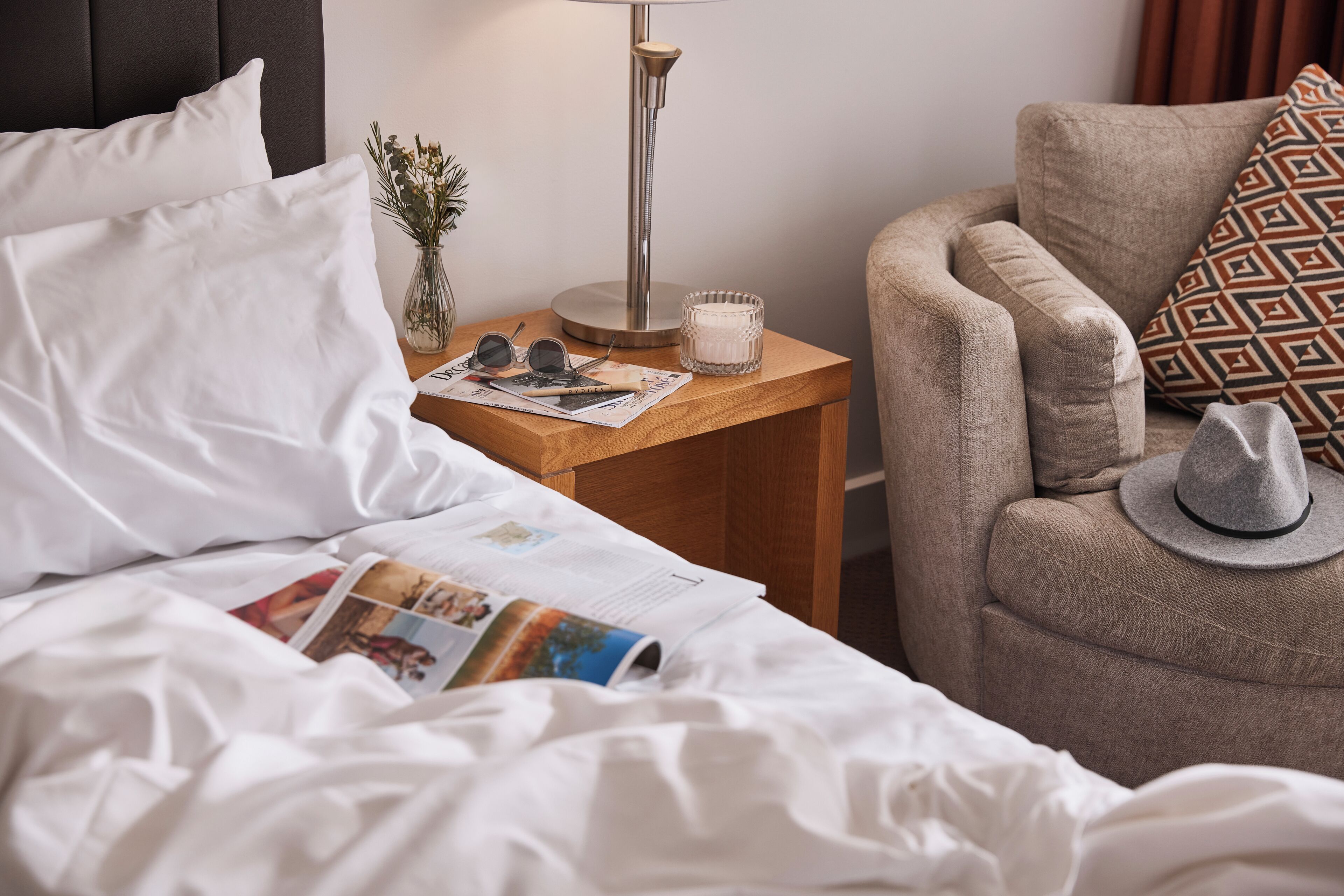 A neatly made bed with white linens, a bedside table with a lamp, a magazine, and a vase with flowers.