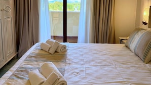 A hotel room with a bed, two rolled towels, a nightstand, a lamp, and a view of greenery through the window. - Hotel Sa Pedra (Olbia)