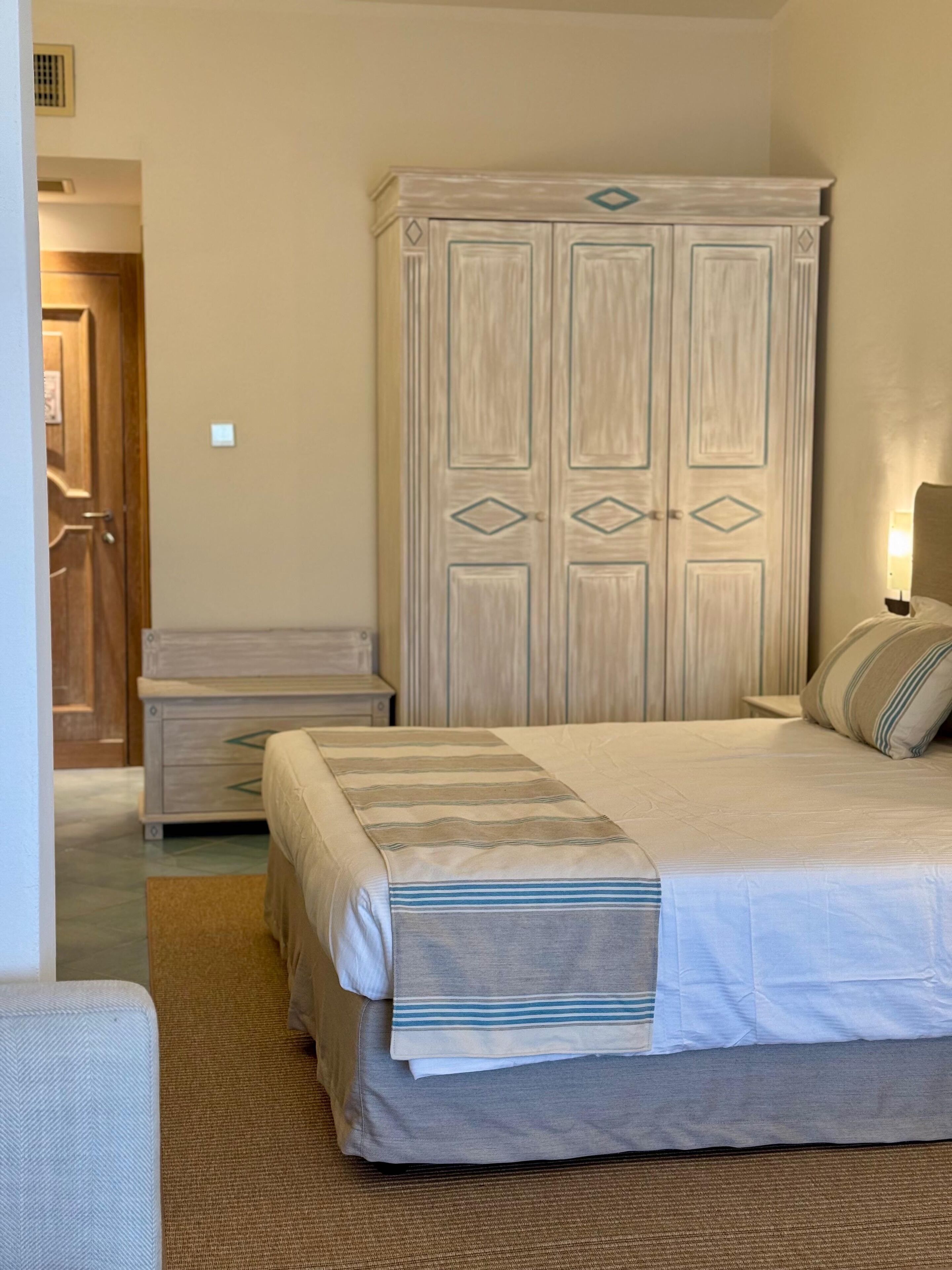 A bedroom with a large bed, a nightstand, a wardrobe, and a door leading to another room.