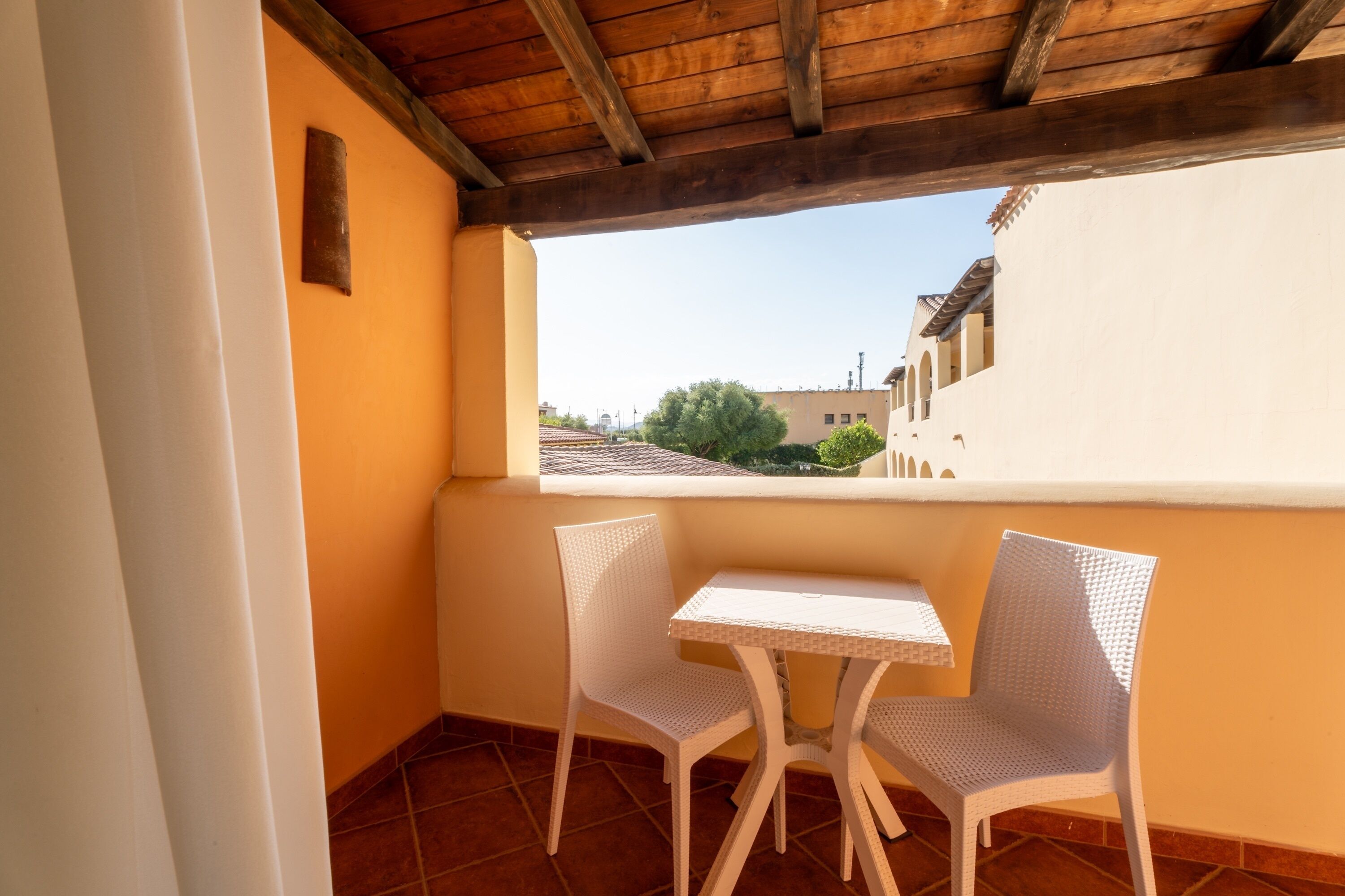 Un balcón con dos sillones blancos de mimbre y una mesita, con vistas a una zona residencial con edificios y árboles.