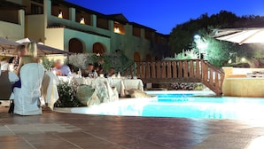 A poolside dining area with tables and chairs, a wooden bridge, and a building with arched windows in the background. - Hotel Sa Pedra (Olbia)