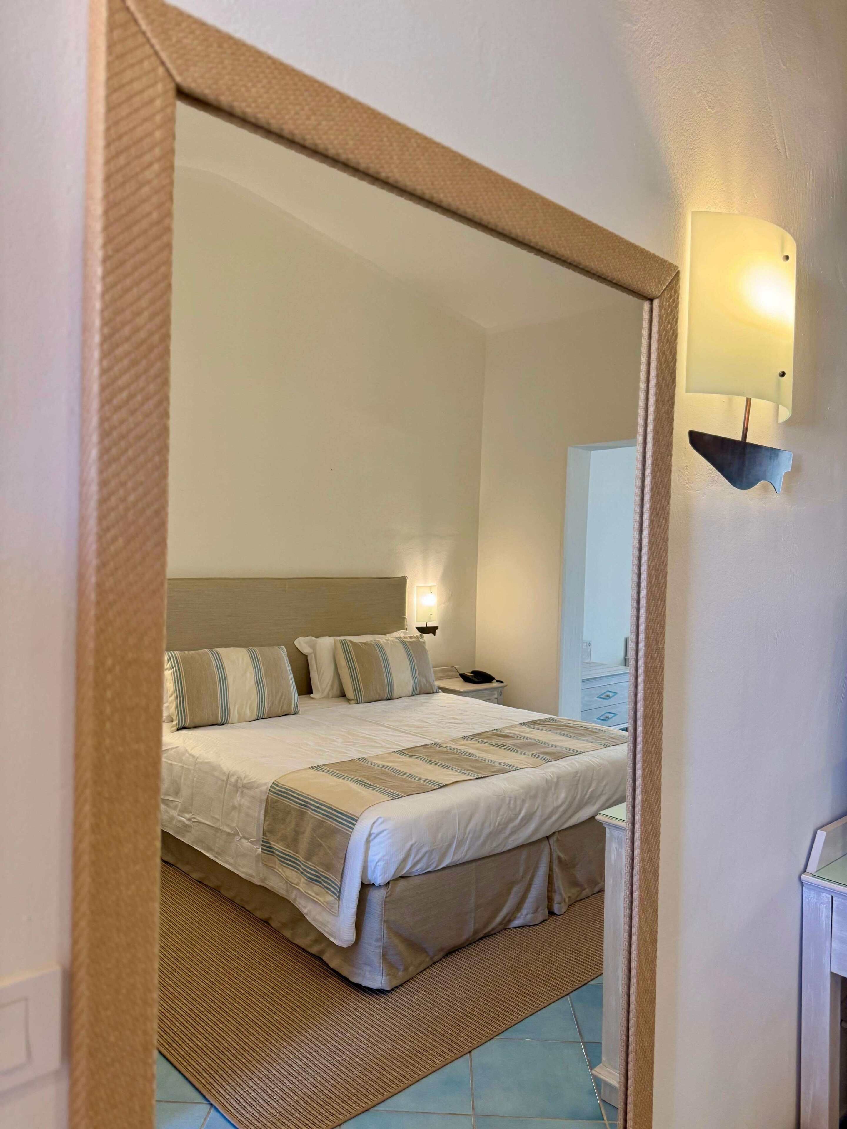 A hotel room with a bed, bedside table, lamp, and a mirror reflecting the room.
