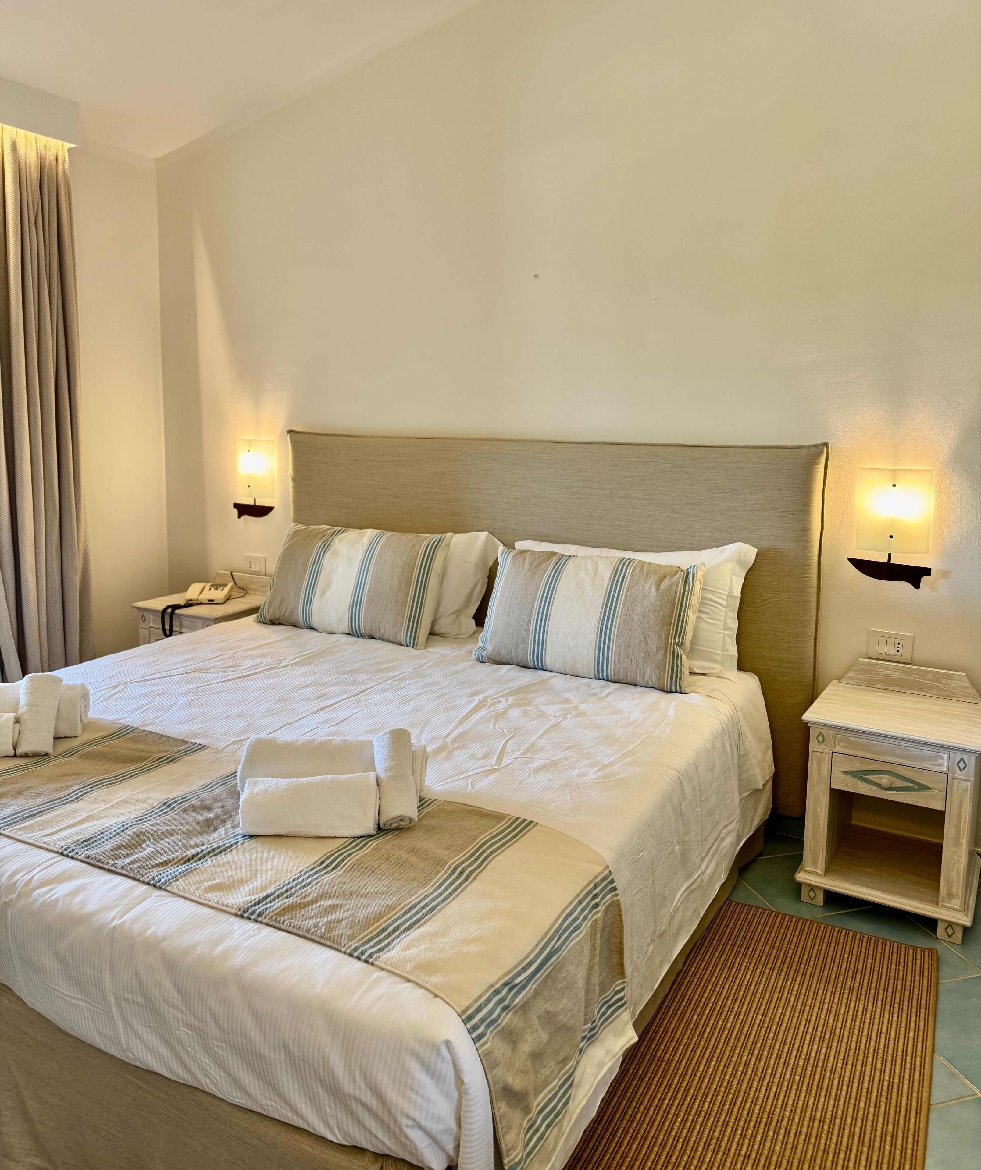 Una cama bien hecha con ropa de cama a rayas, dos mesitas de noche con lámparas y un teléfono junto a la cama.
