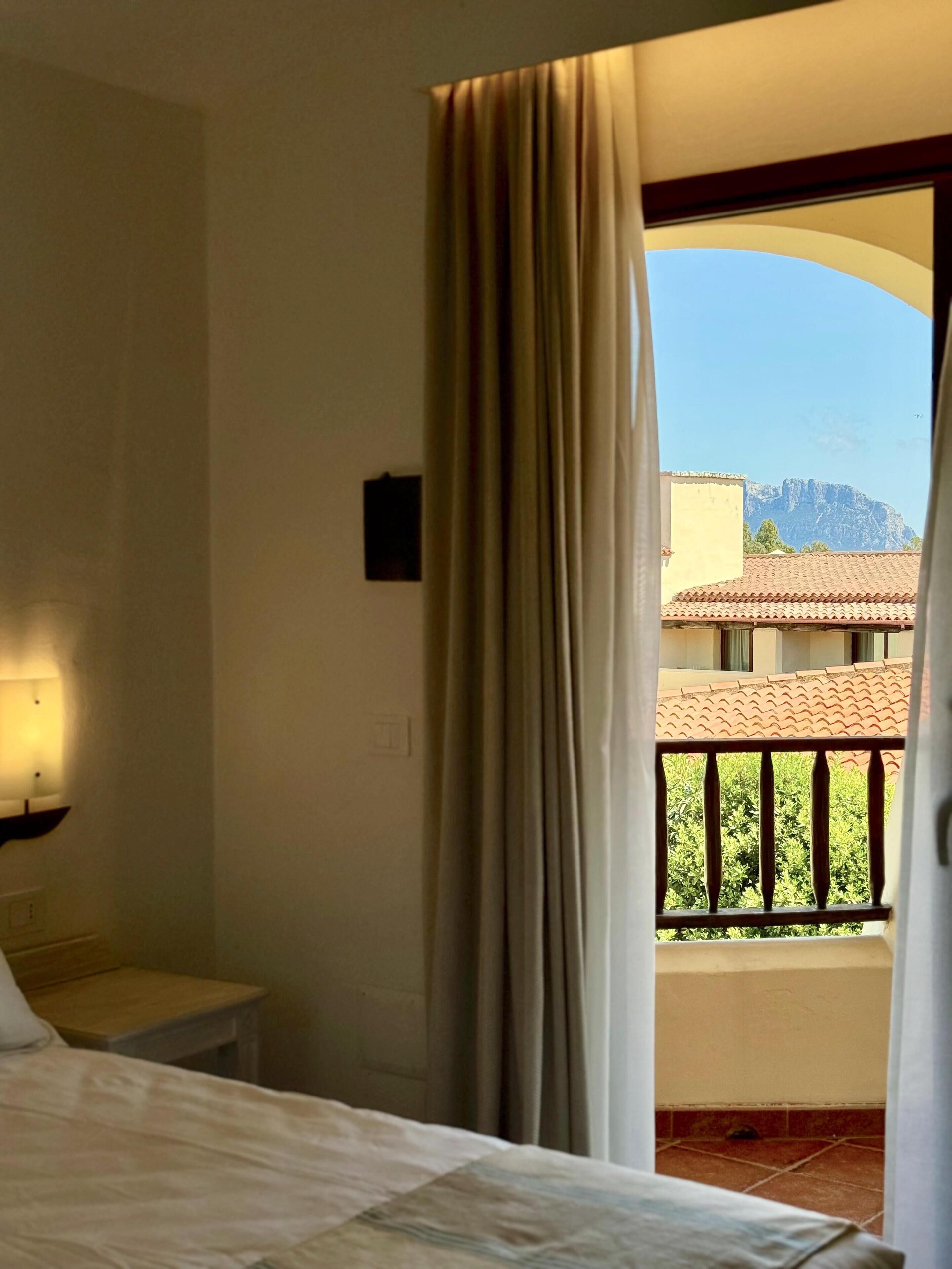 A hotel room with a bed, a nightstand, a lamp, a window with curtains, and a view of a building and mountains.