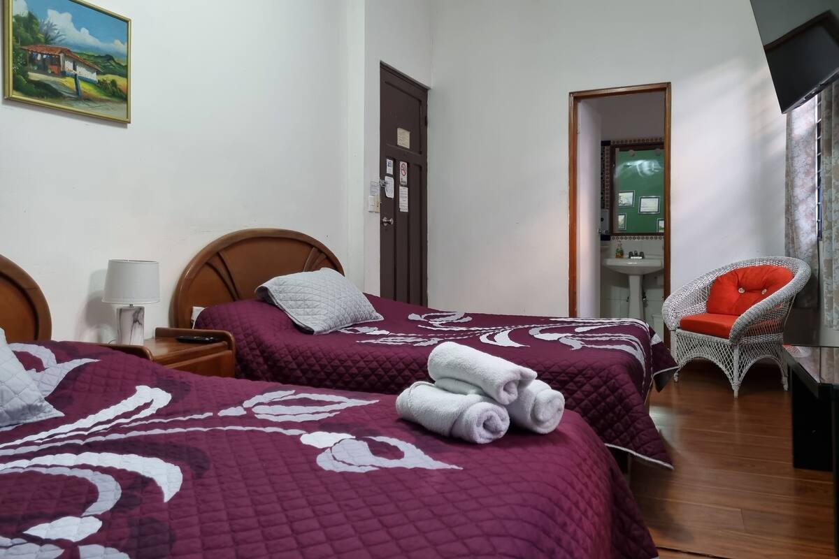 A hotel room with two beds, a chair, a painting, and a bathroom visible through an open door.