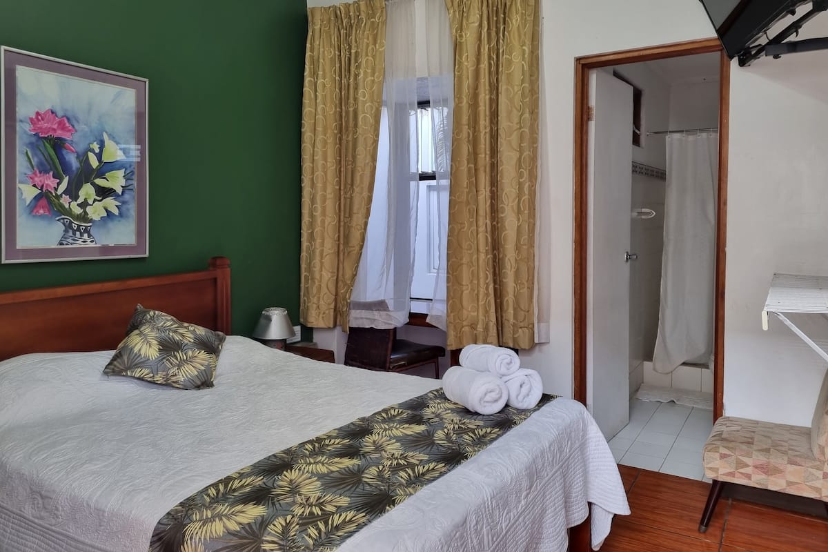 A neatly made bed with a floral bedspread, a painting of flowers on the wall, and a bathroom visible through a doorway.