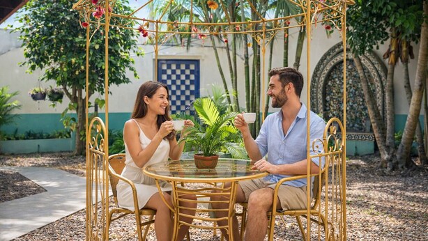 A couple seated at a round table with a potted plant, under a decorative metal gazebo in an outdoor setting with greenery and architectural details. - Hotel 1492 (San José)
