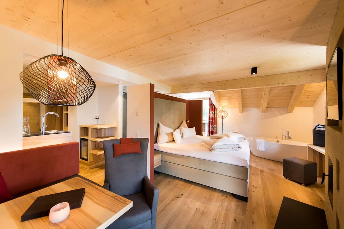 Ein modernes Hotelzimmer mit Holzbalkendecke, einem Bett mit weißer Bettwäsche, einer Sitzecke mit Stuhl und Tisch sowie einer kleinen Küchenzeile.