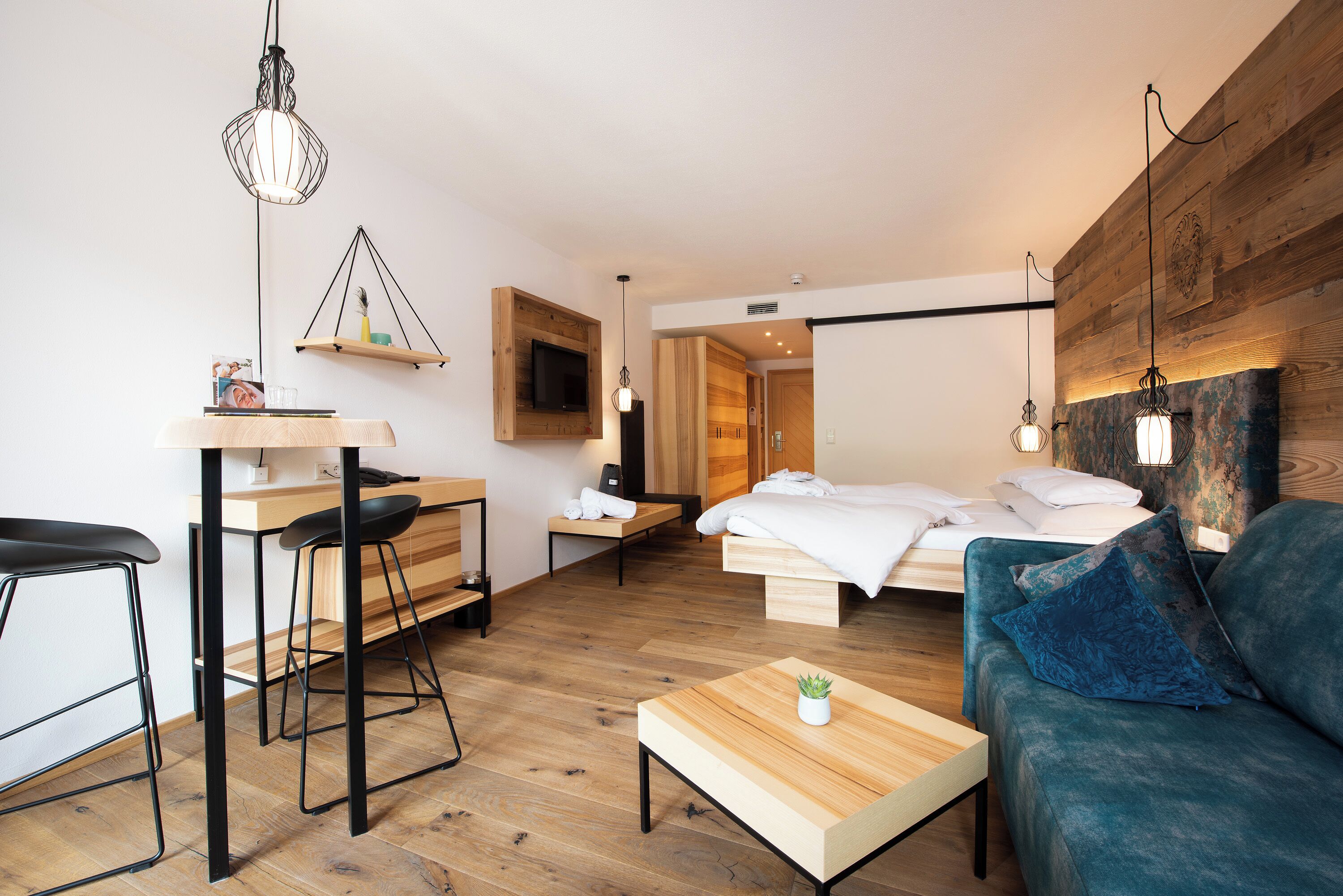 Una habitación de hotel moderna con piso de madera, una cama con sábanas blancas, un sofá azul, una mesita de centro de madera y un escritorio con una silla.