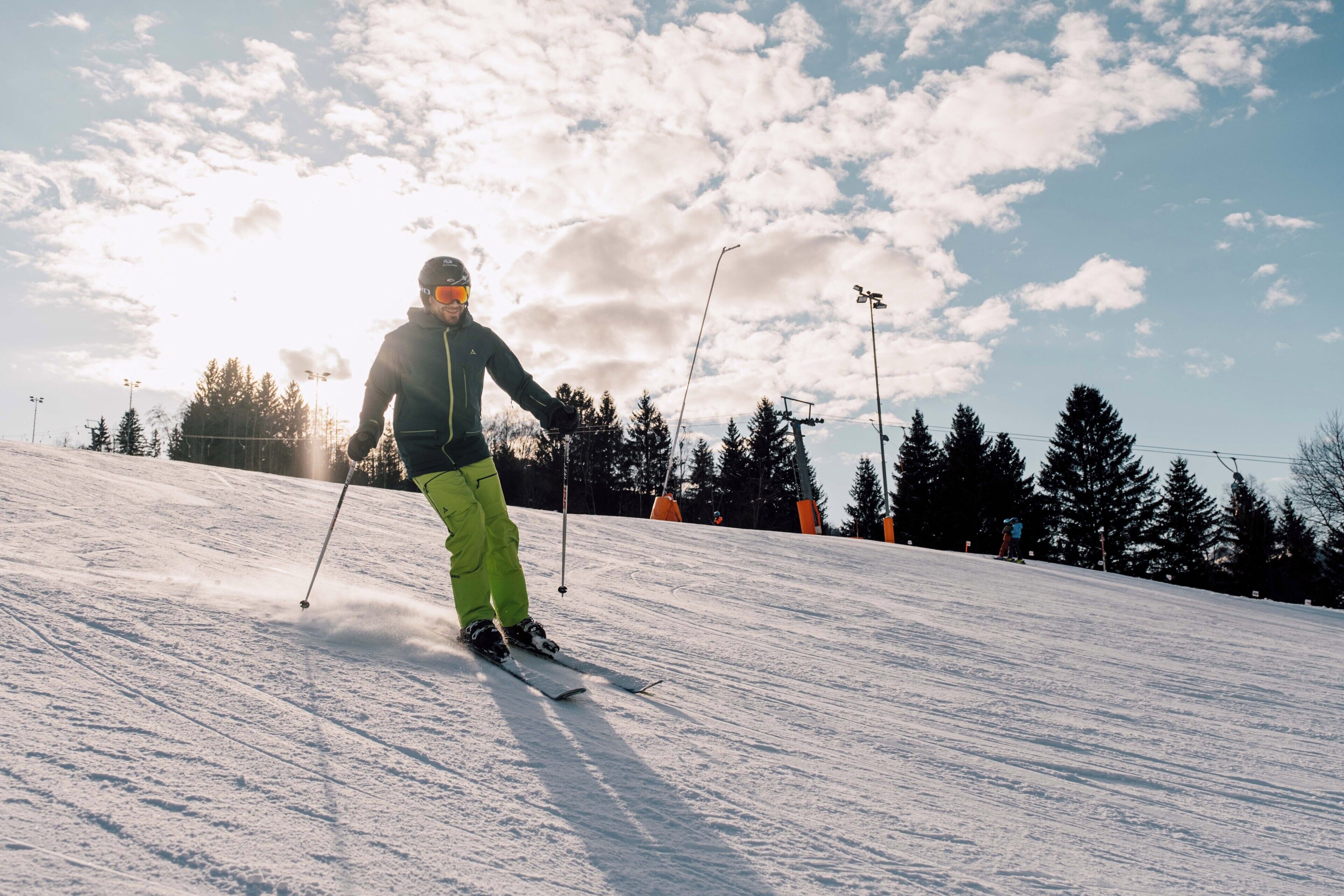 A person skiing down a snowy slope with trees and ski lifts in the background.