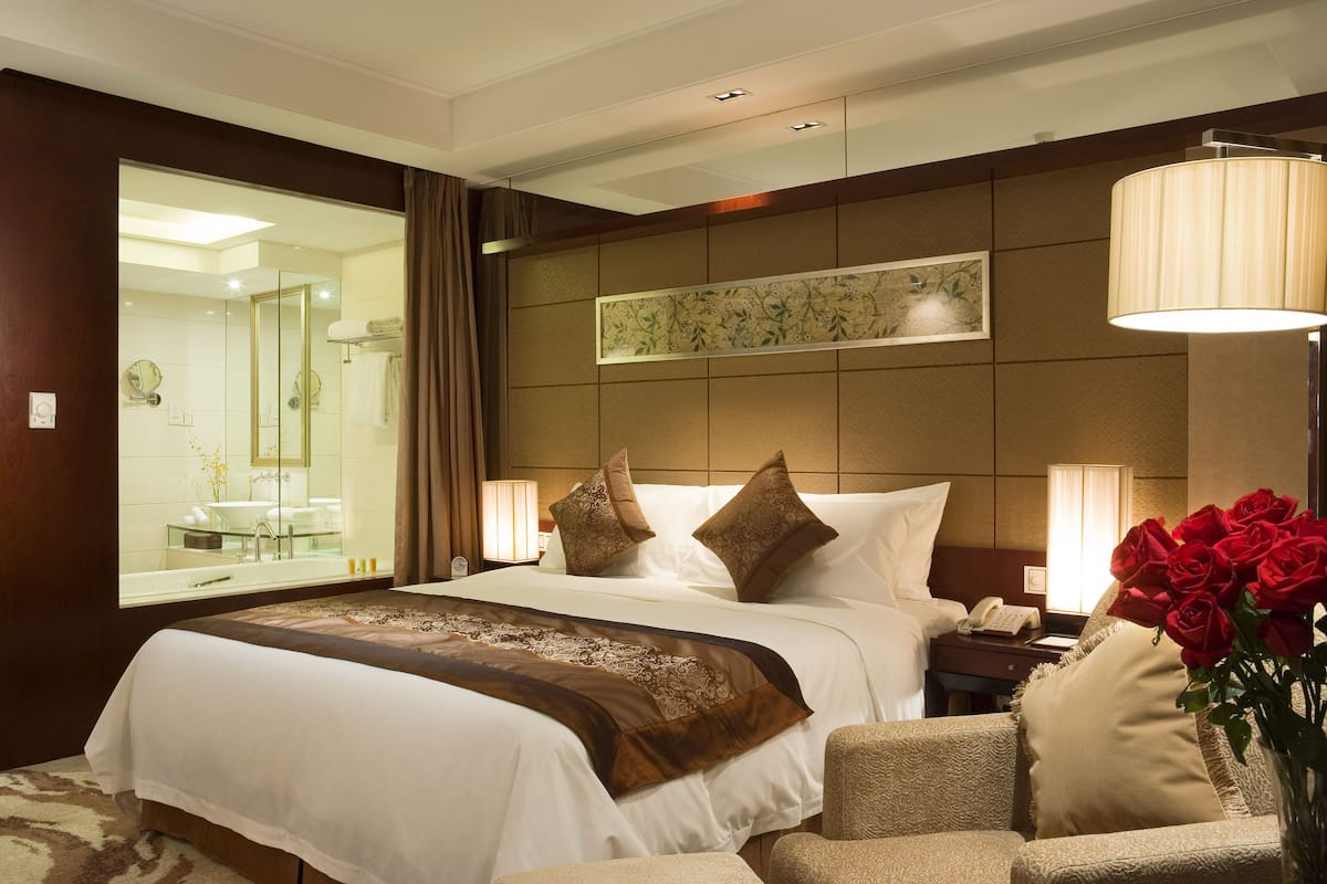 Une chambre d’hôtel avec un grand lit, un coin salon avec un fauteuil, une table de chevet, une lampe et un vase avec des fleurs.