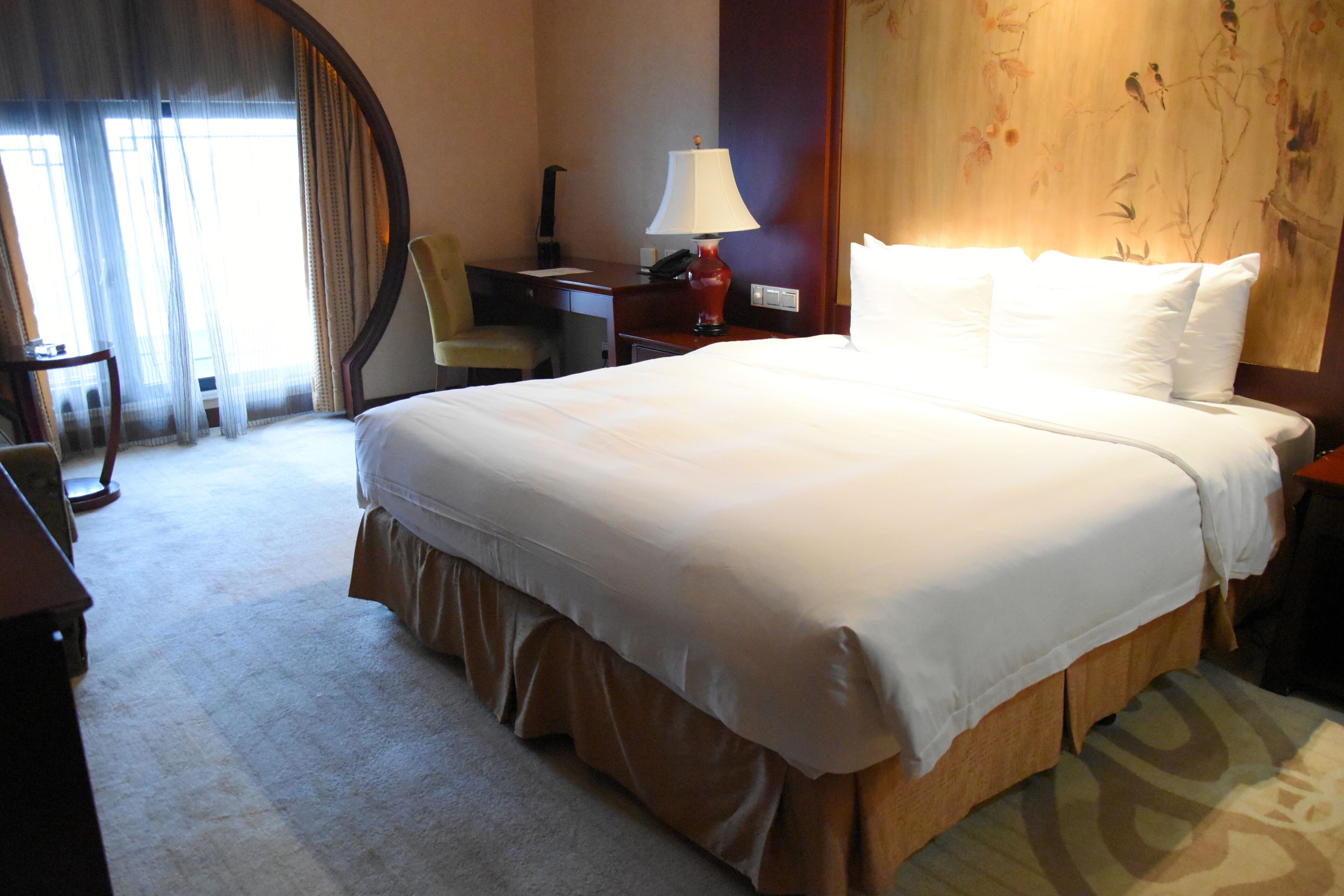 A hotel room with a large bed, a desk, a chair, a lamp, and a painting on the wall.