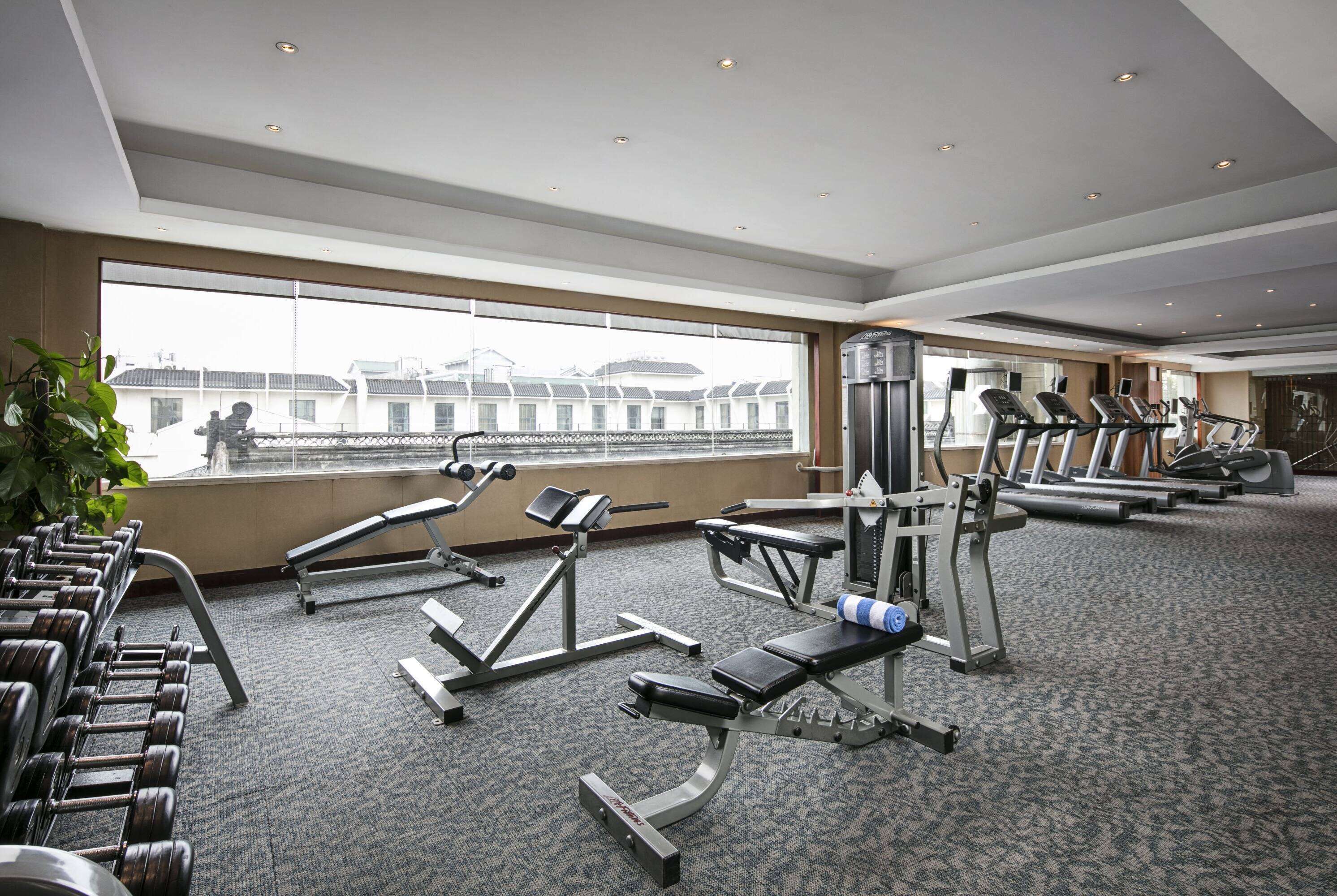 A gym with various exercise equipment, large windows, and a view of buildings.
