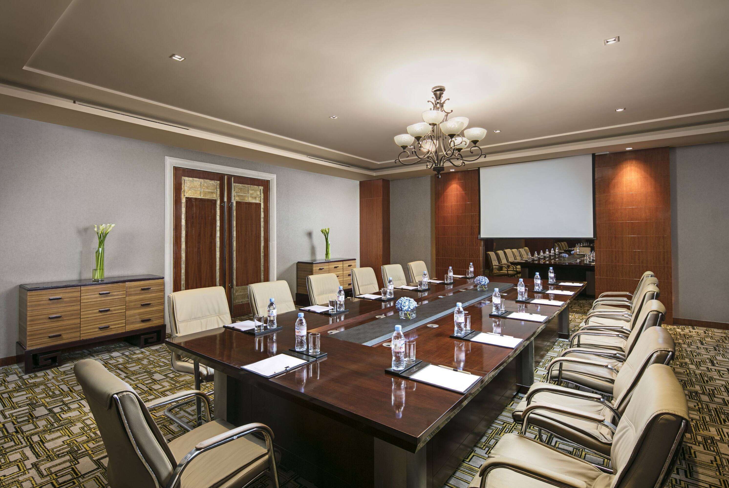 A conference room with a long table, multiple chairs, a chandelier, a wooden cabinet, and a patterned carpet.