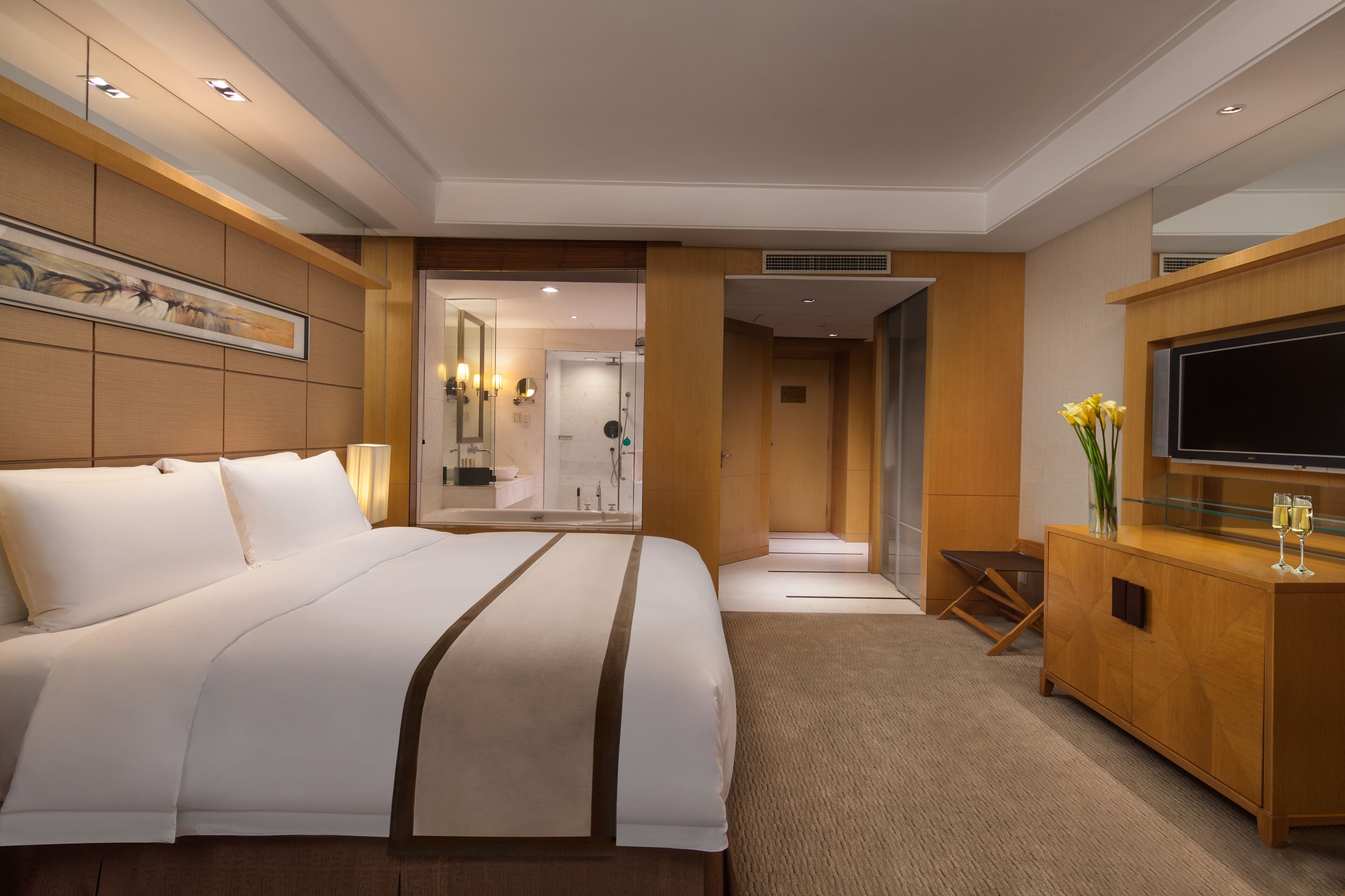 A modern hotel room with a large bed, a wooden console table with a flat-screen TV, and a bathroom visible through an open door.