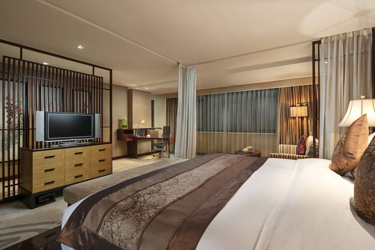 Une chambre d’hôtel moderne avec un grand lit, un téléviseur posé sur un meuble en bois, un bureau avec une chaise et une grande fenêtre avec des rideaux.