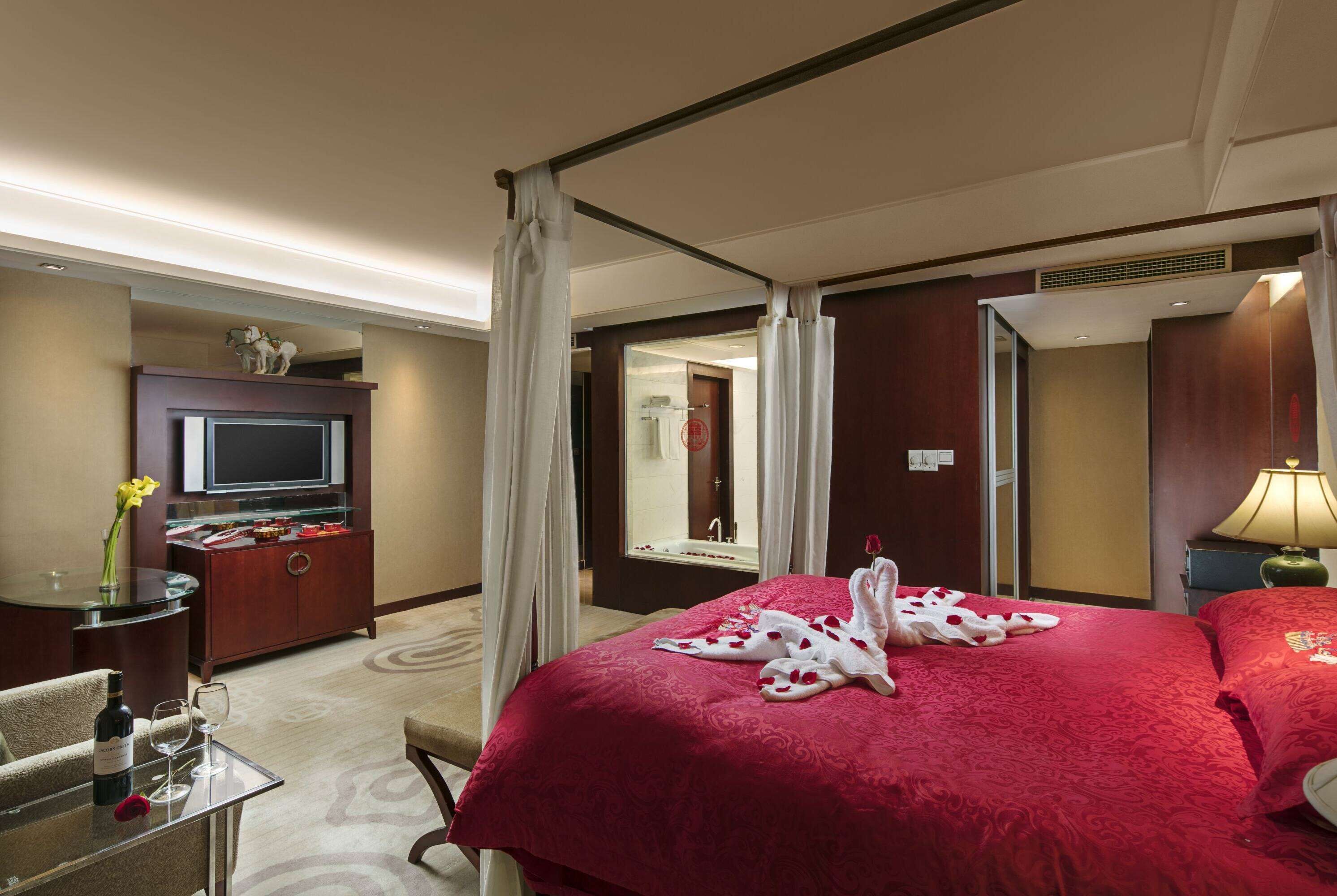 A hotel room with a large bed, a TV, a minibar, a desk with a glass top, and a bathroom visible through an open door.