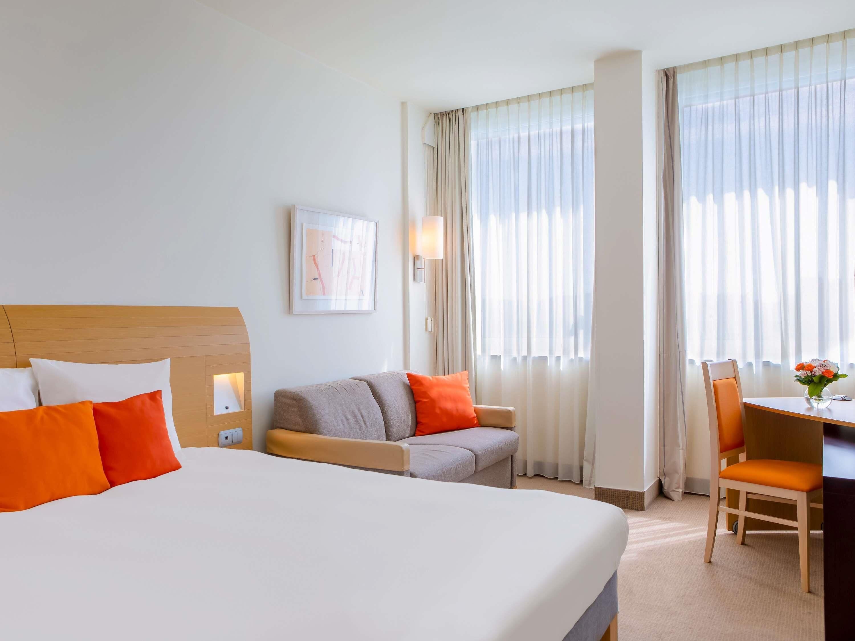 A hotel room with a large bed, a sofa, a desk, and two windows with curtains.