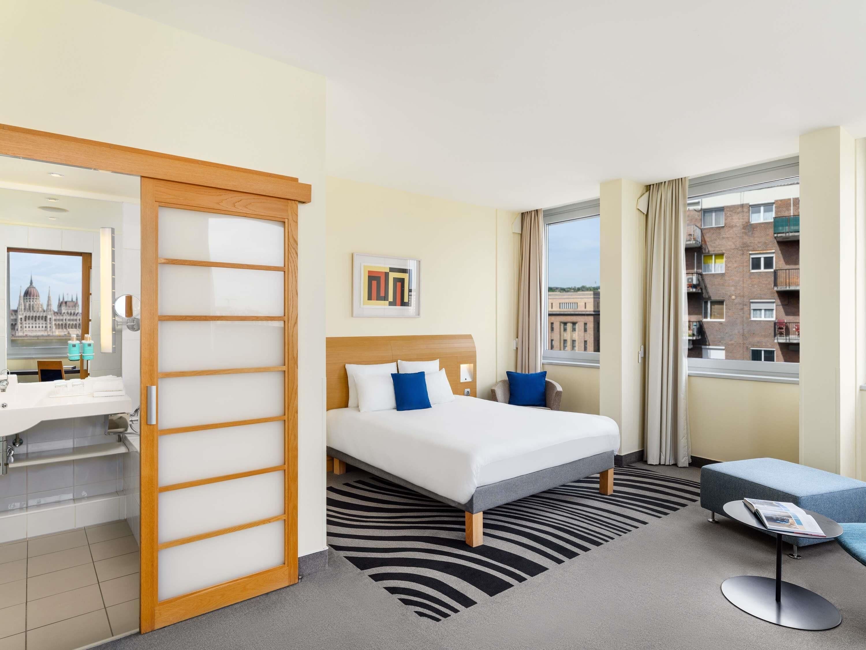 A hotel room with a bed, a desk, a mirror, a window with a city view, and a zebra-patterned rug.